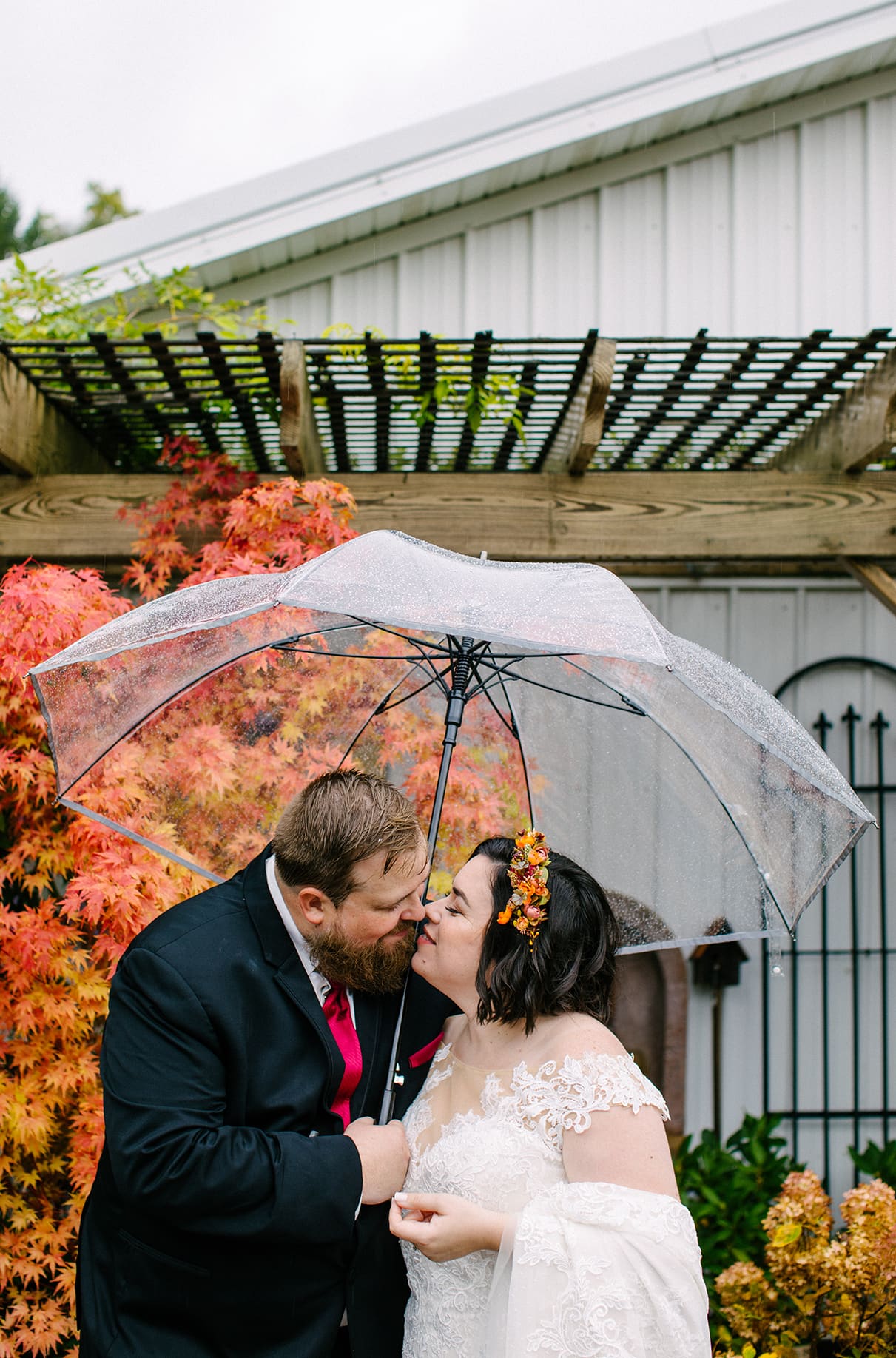 Bride and Groom stand under clear umbrella in front of tree with orange leaves during the fall at Carol Watson Greenhouse in Lafayette NY