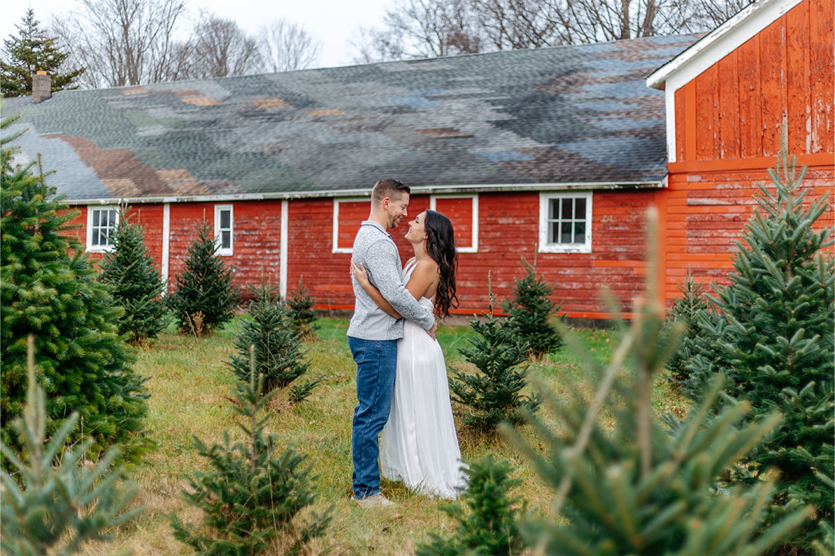 Couple looks at each other lovingly while standing in front of red barn and Christmas trees at Sipples Farm in Bainbridge NY