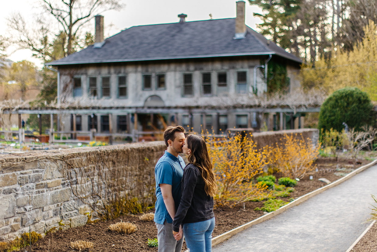 Couple kisses in front of historic home for engagement photos at Cornell Botanic Gardens in Ithaca NY