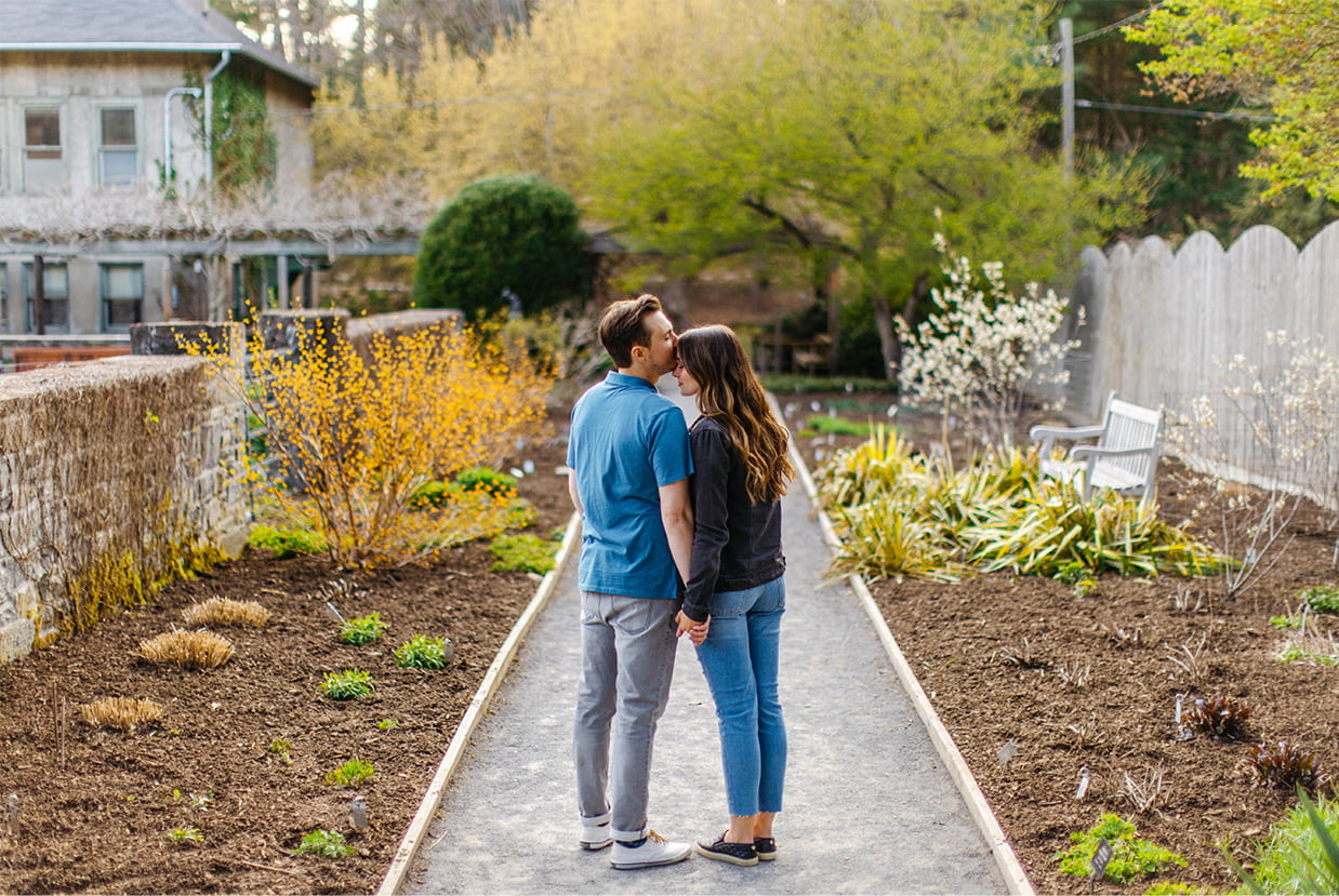 couple holding hands. man kisses womans forehead as they stand on garden path at Cornell Botanic Gardens in Ithaca NY