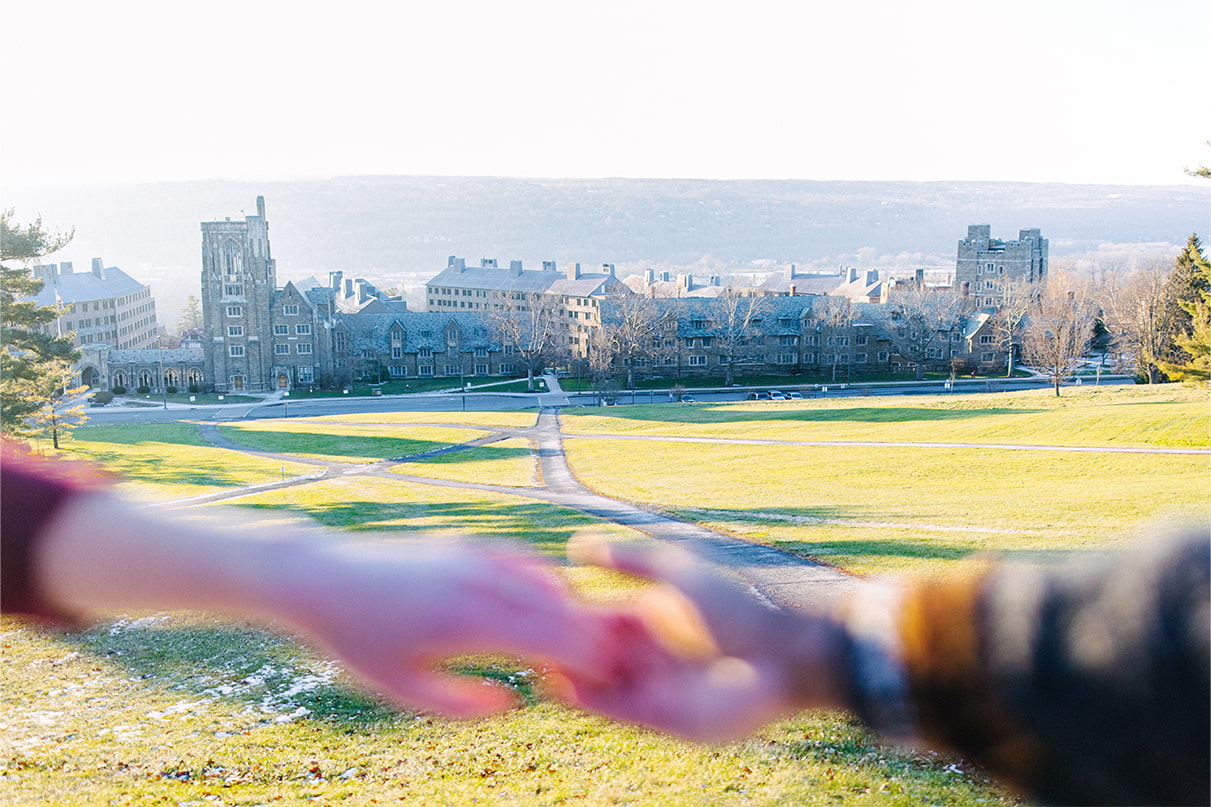couple holds hands out of focus in front of camera, with Cornell University campus in focus behind them