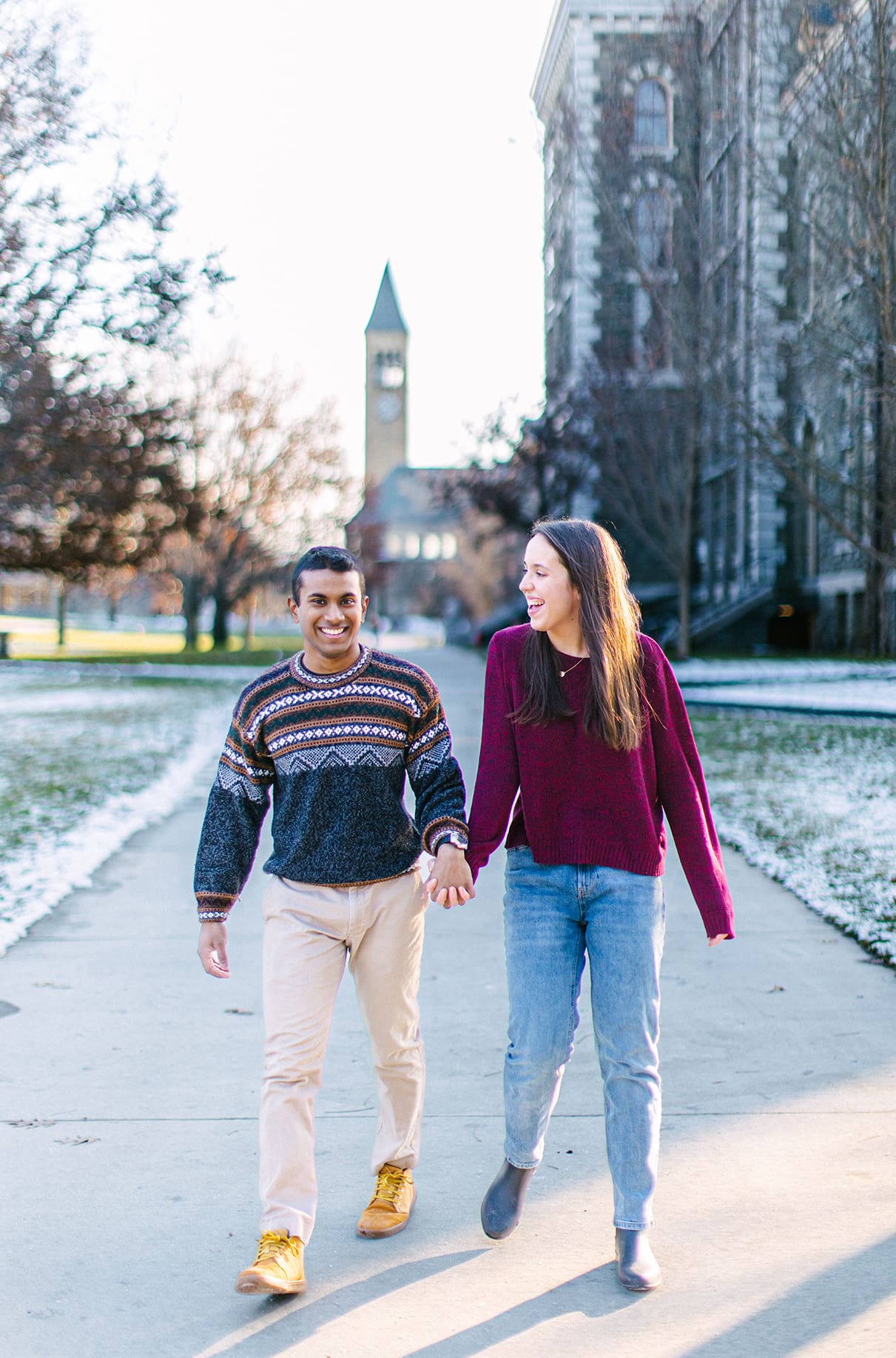 Couple holds hands while walking and laughing in front of the clock tower at the Cornell University Campus in Ithaca, NY
