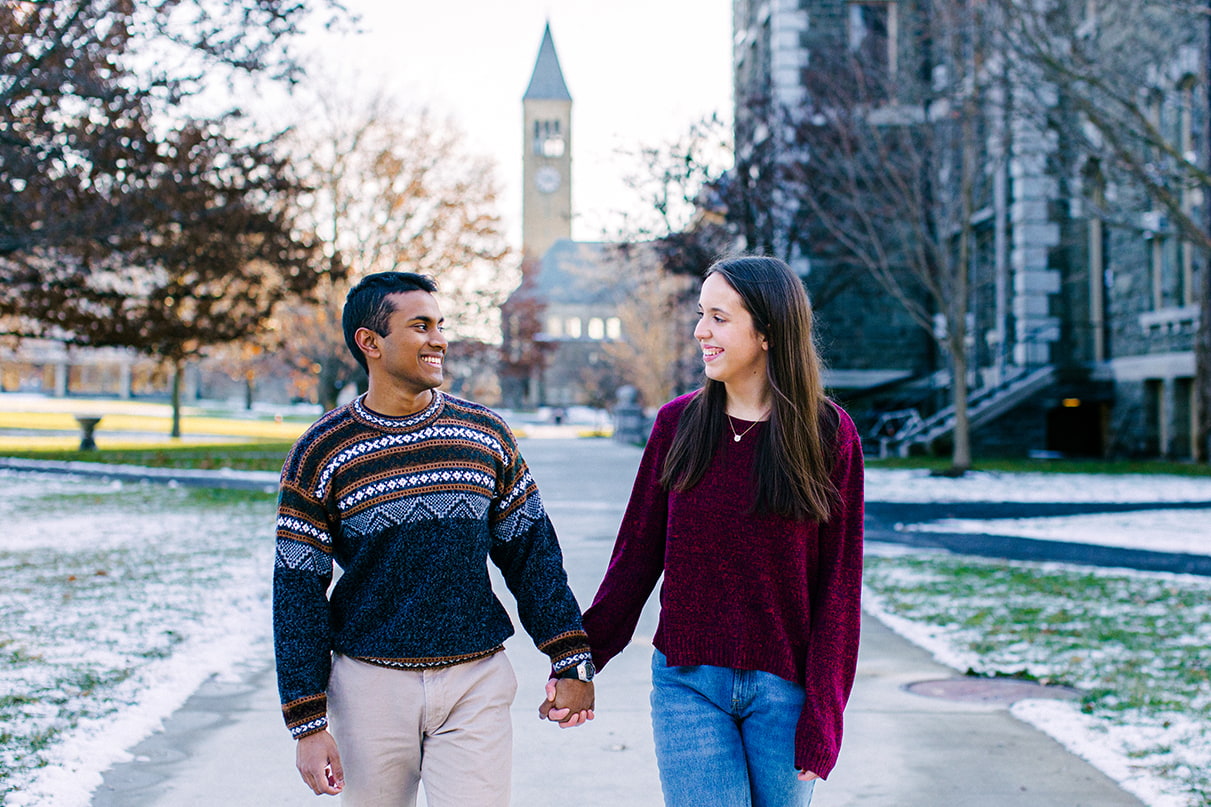 Couple smiles and holds hands in front of the Cornell clock tower in Ithaca, NY
