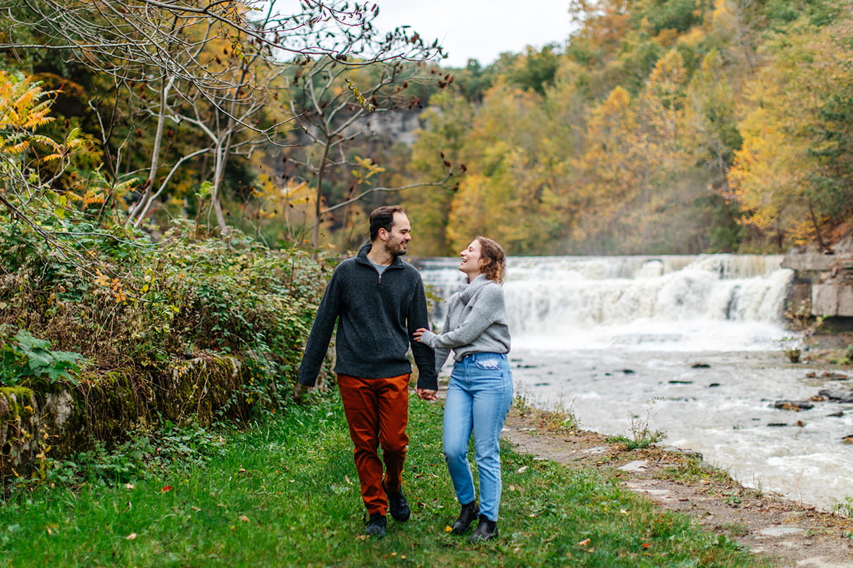 Couple holds hands and walks in front of falls at Taughannock Falls State Park