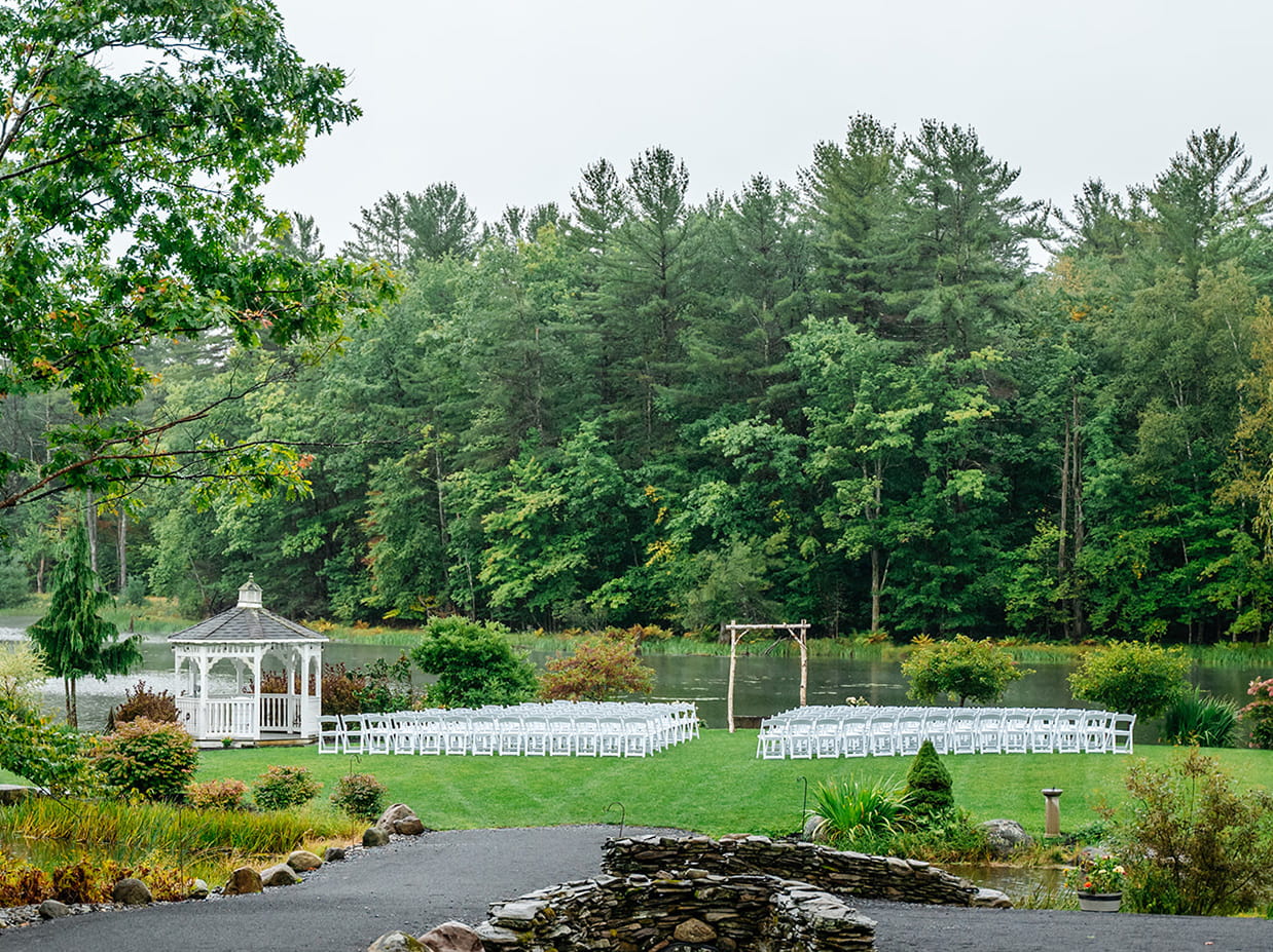 Wooded lakeside wedding ceremony space set up with white chairs, a birch arbor, and a white gazebo