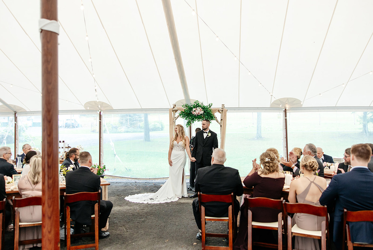 Bride and groom look to their guests during their tented wedding ceremony at The Rosemary at Spano Lake in Schenevus, NY