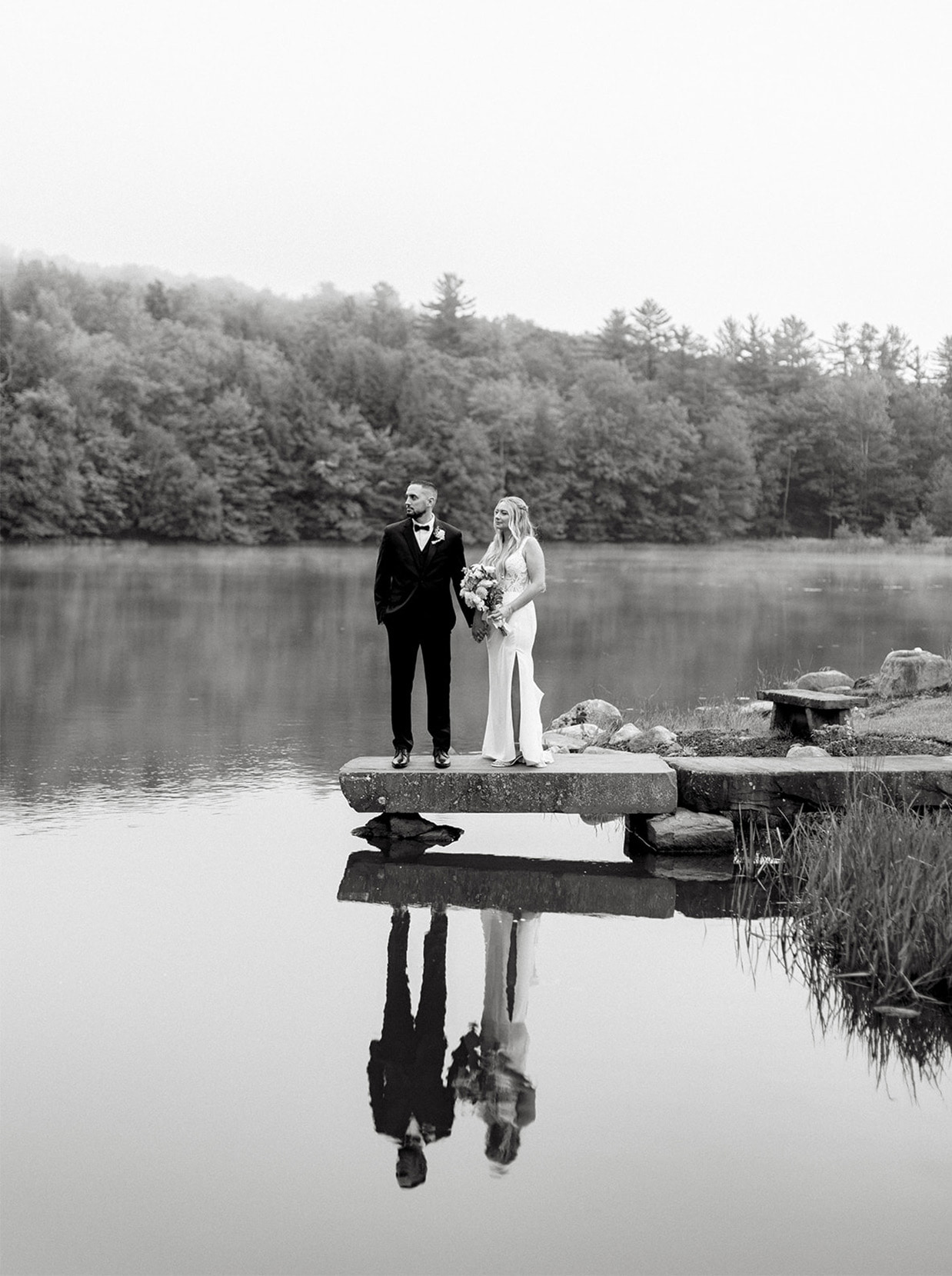 Black and white photograph of a bride and groom on a stone dock at The Rosemary at Spano Lake. They are reflected in the water below them