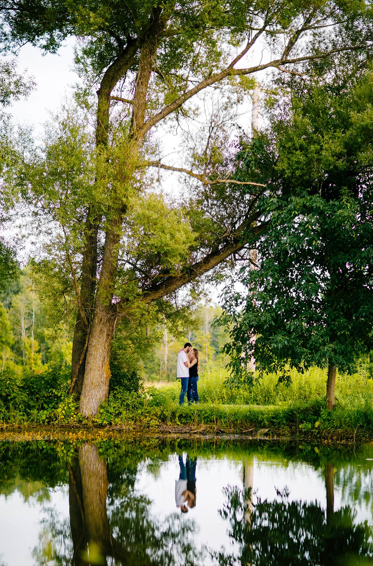 couple embraces between trees next to pond and their reflection is mirrored in the water Albany NY