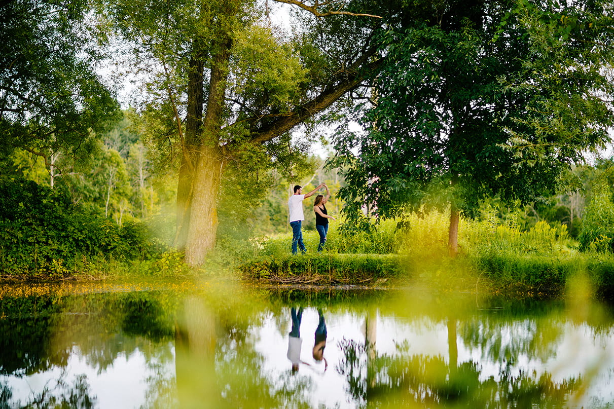 Couple dances and man twirls woman between trees next to pond and their reflection is mirrored in the water