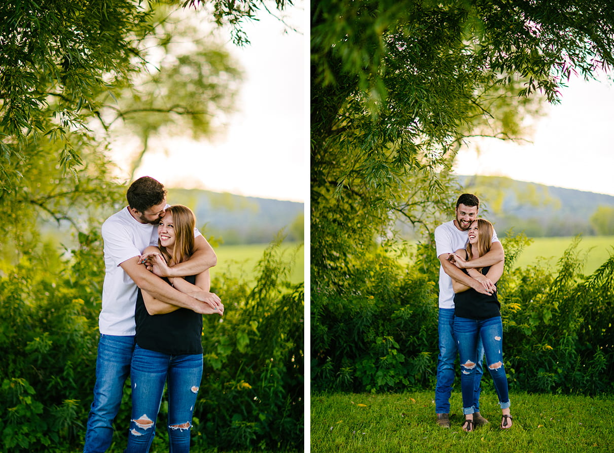 Couple stands in front of bushes. Man stands behind woman and hugs her while they both smile and laugh