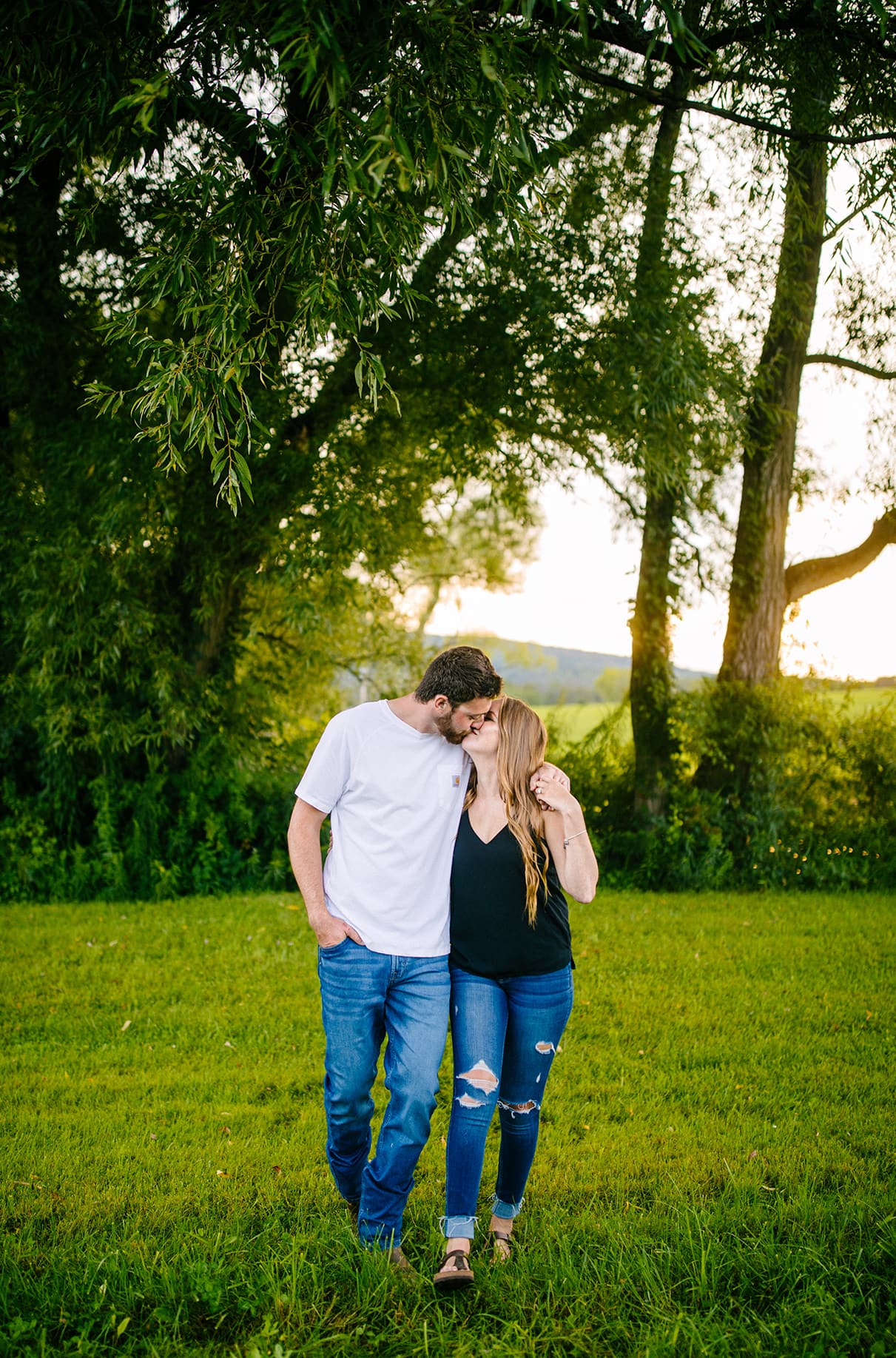 Couple walks towards camera during golden hour with trees and shining sun behind them. They kiss. Albany NY