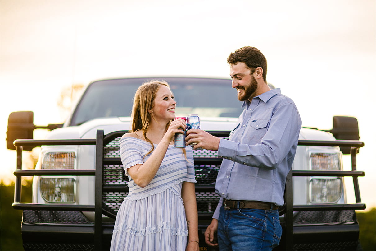couple toasts with beer cans in front of Ford truck while smiling
