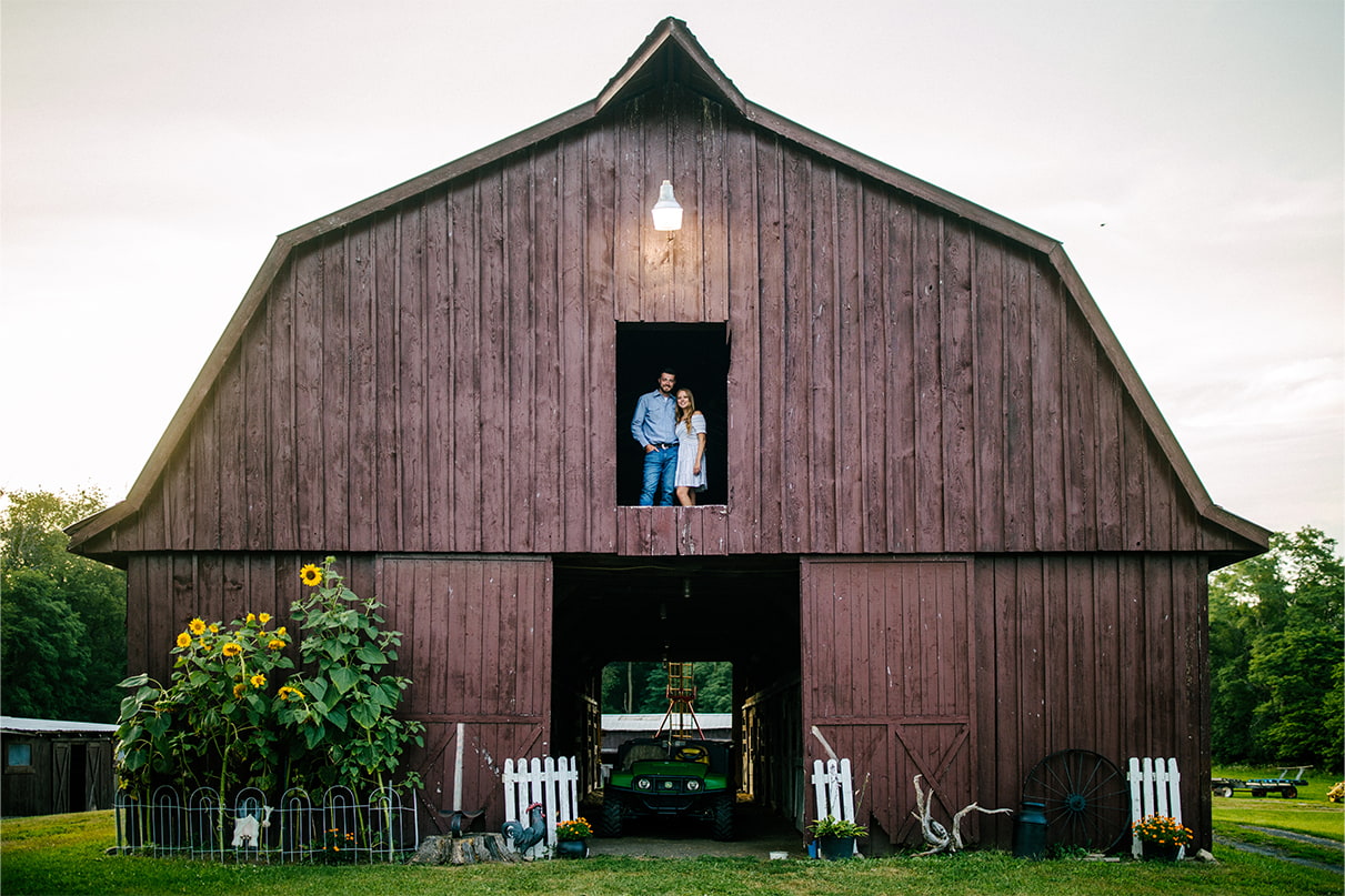 couple stands framed in barn window on second story of barn. There are sunflowers planted outside the barn.