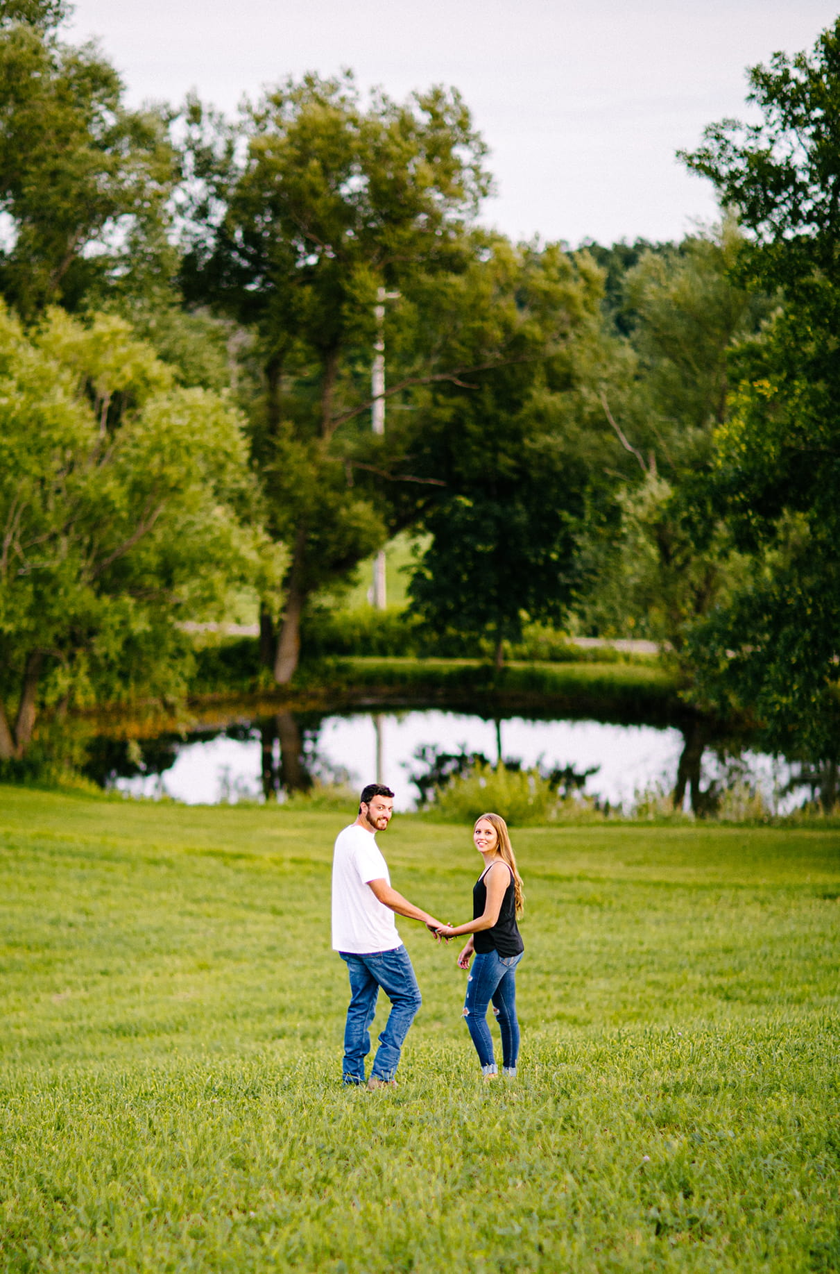 Couple holding hands and walking down hill in front of pond while looking back at the camera