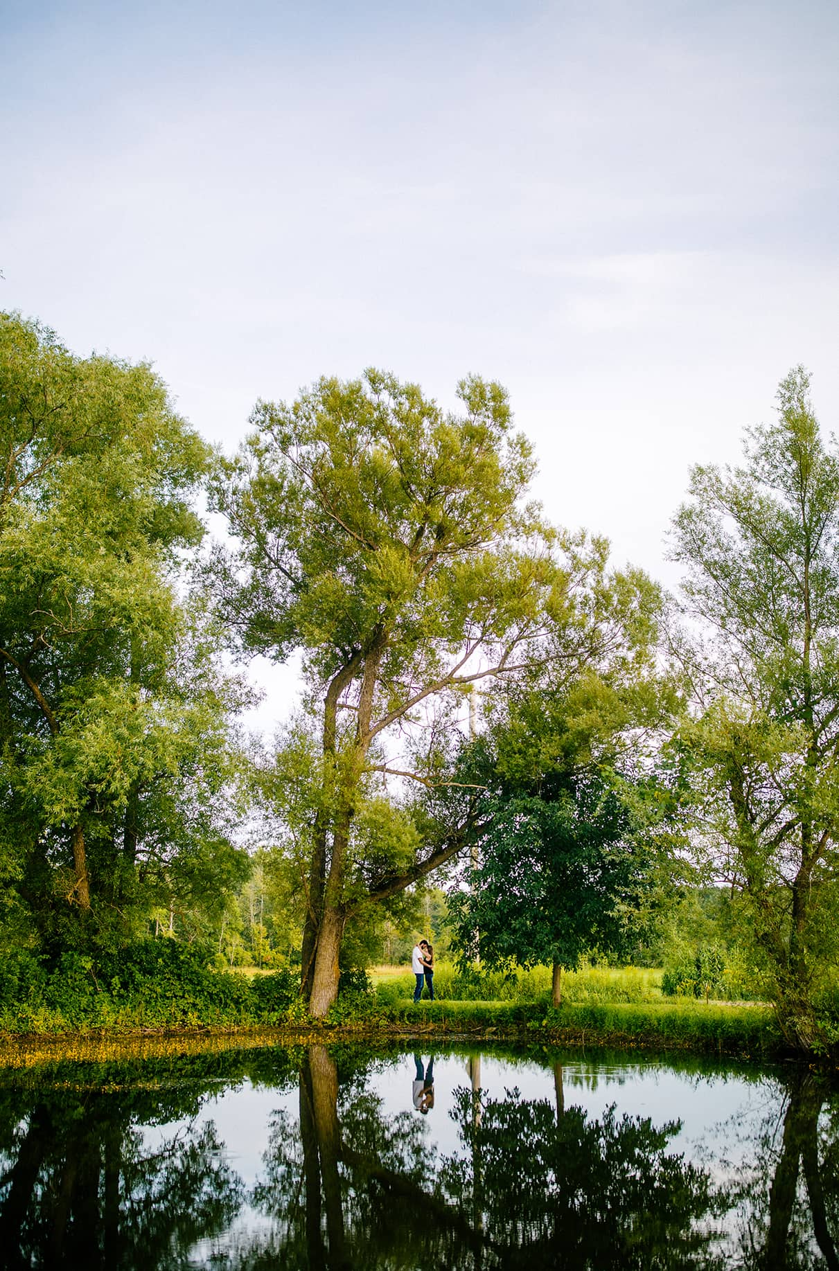 Man and woman stand between trees behind a pond and their reflection is mirrored in the pond Albany NY