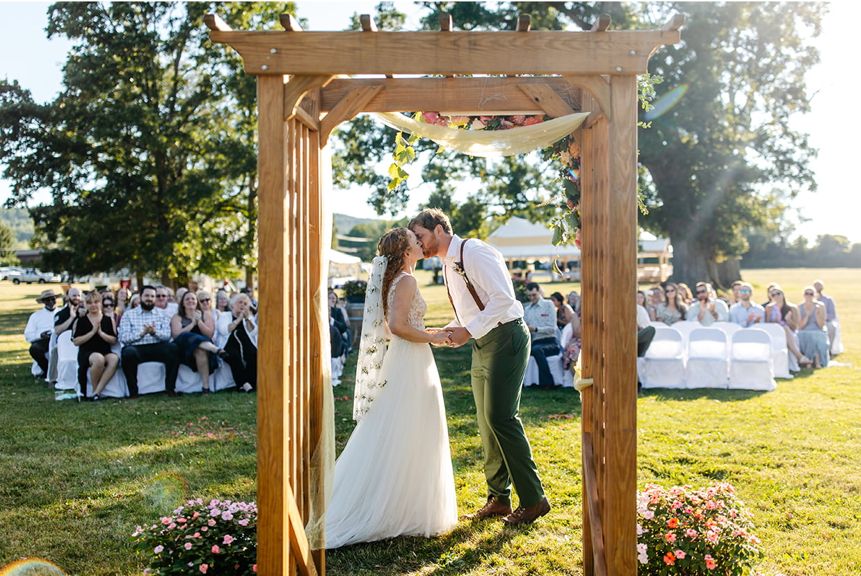 bride and groom share their first kiss while guests clap during their sunny wedding ceremony at Apple Blossom Acres in Freeville NY