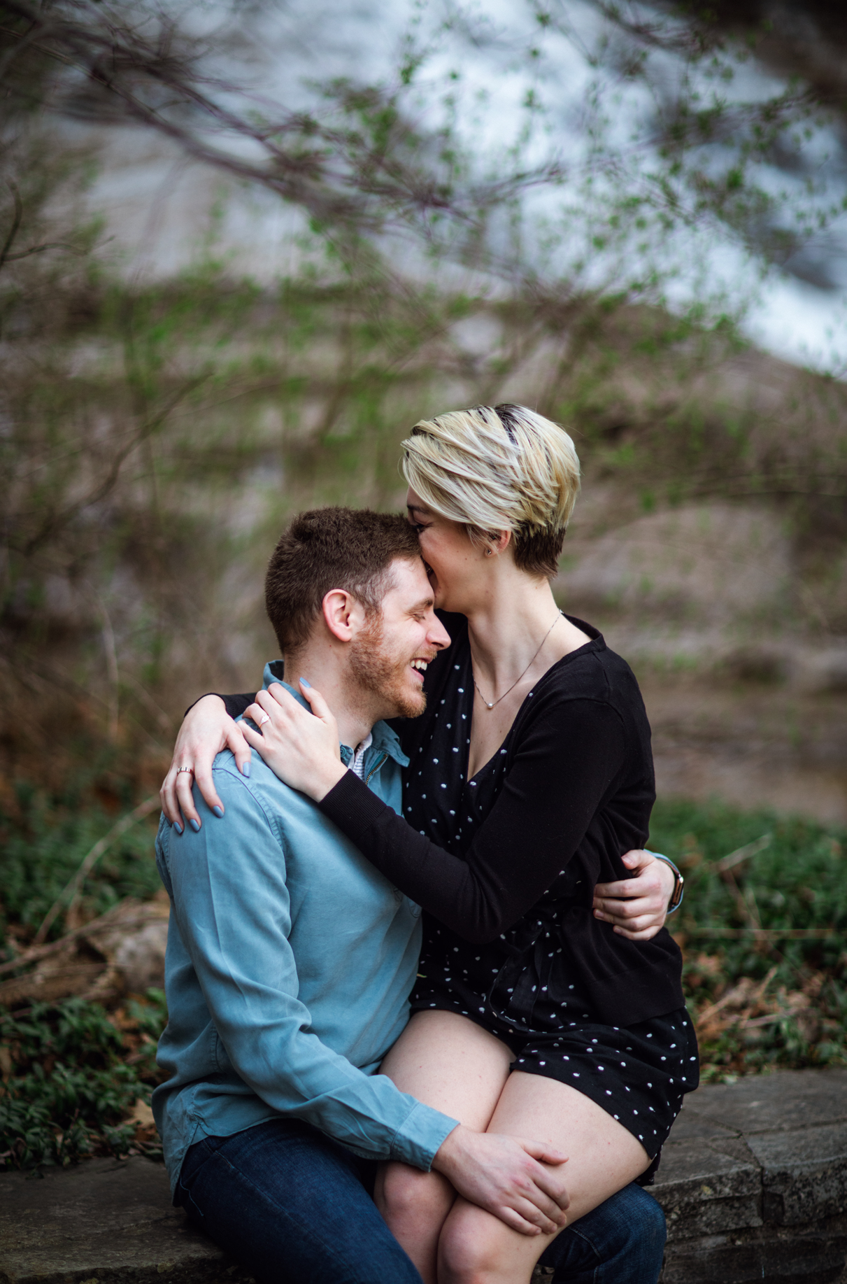 Woman sits on mans lap with arms around him and laughs engagement session