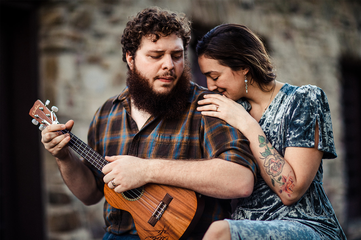 Man plays ukulele while woman rests on his shoulder during engagement session
