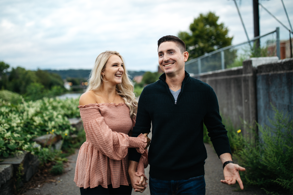 Couple smiles and laughs while holding hands and walking beside river during engagement photos