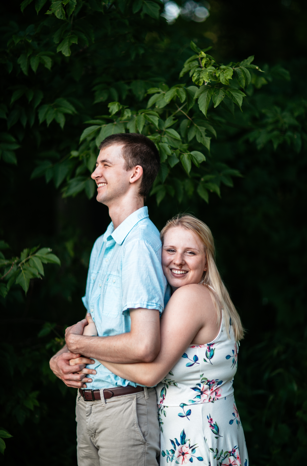 woman hugs man from behind while laughing and standing in front of large bush