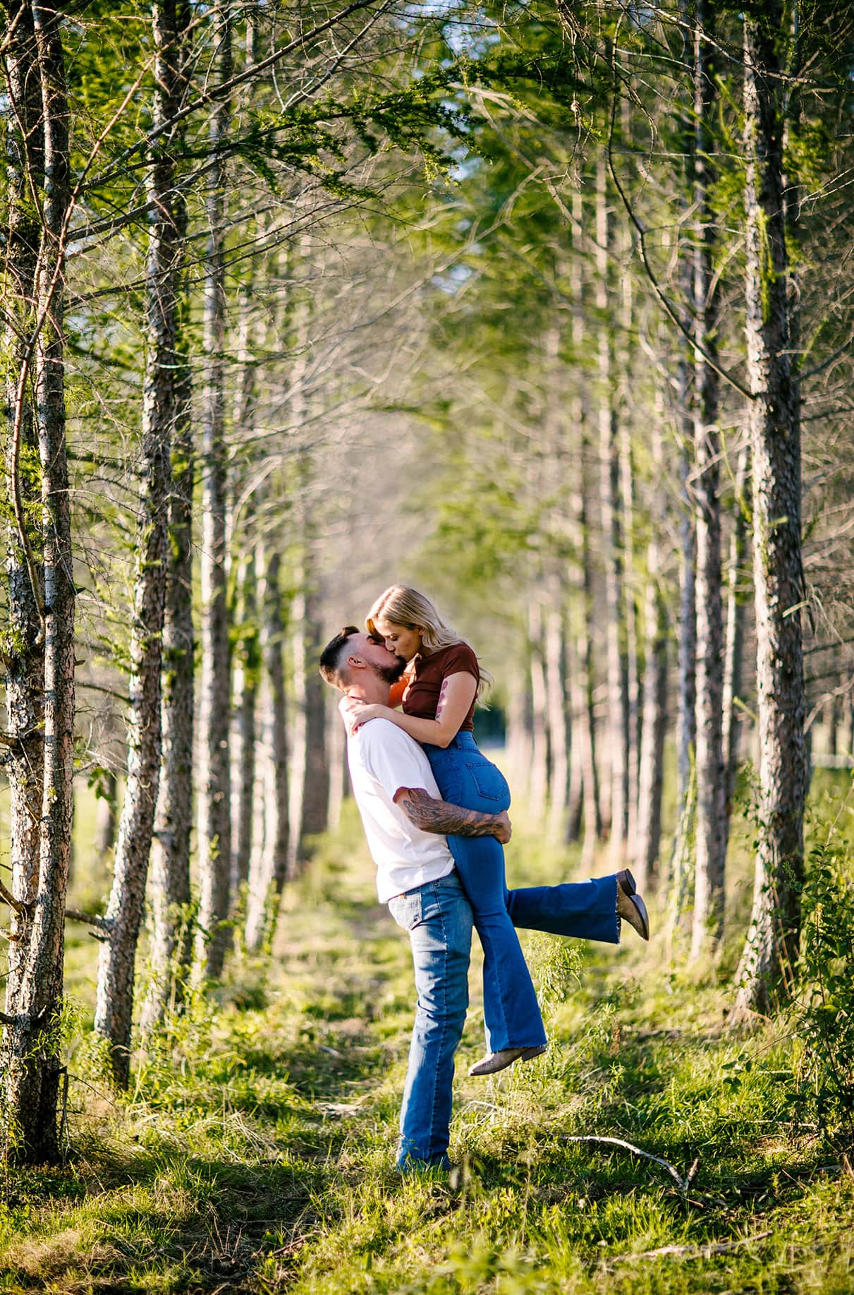 man lifts woman as they kiss in the middle of a line of trees