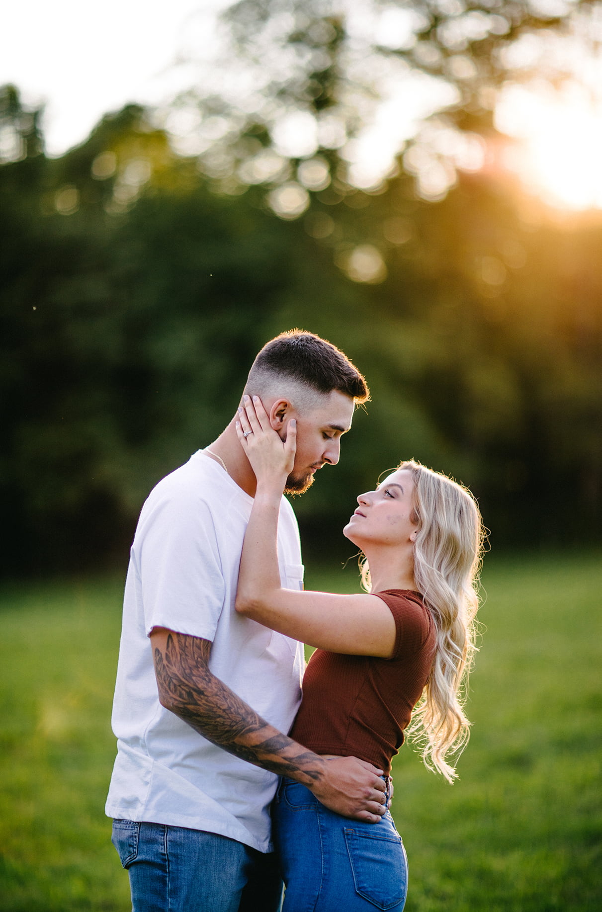 Woman places hand on mans face during golden hour as sun sets behind trees