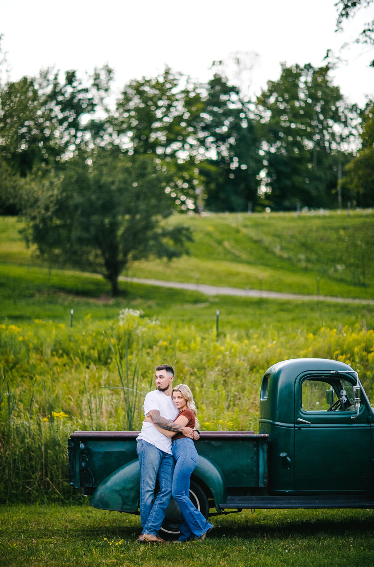 couple embraces while standing in front of a hillside and green vintage chevy truck