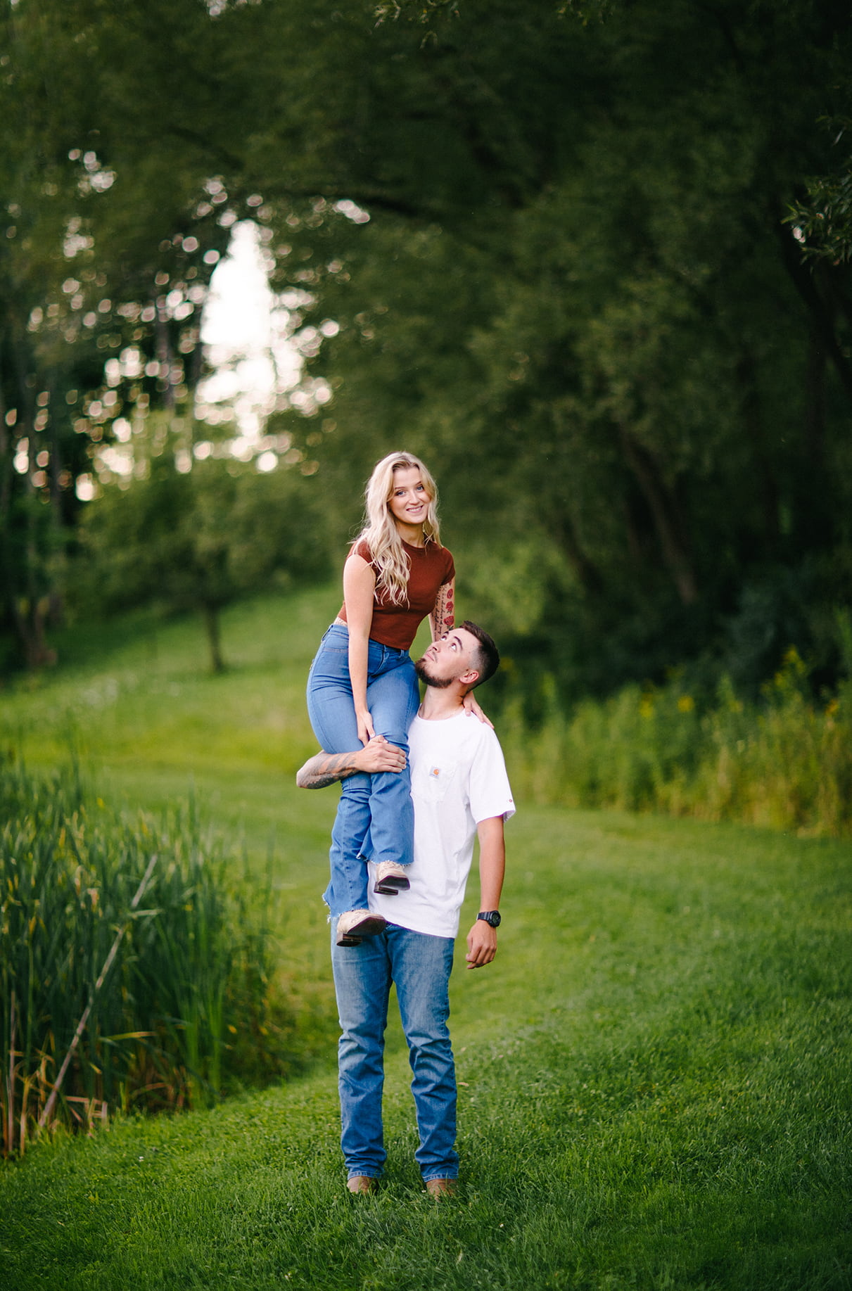 Woman sits on mans shoulders in front of a pond and they look at eachother