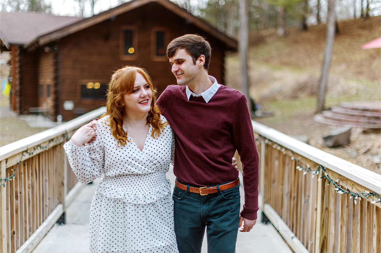 Couples holds hands and smiles while walking across a bridge in the woods