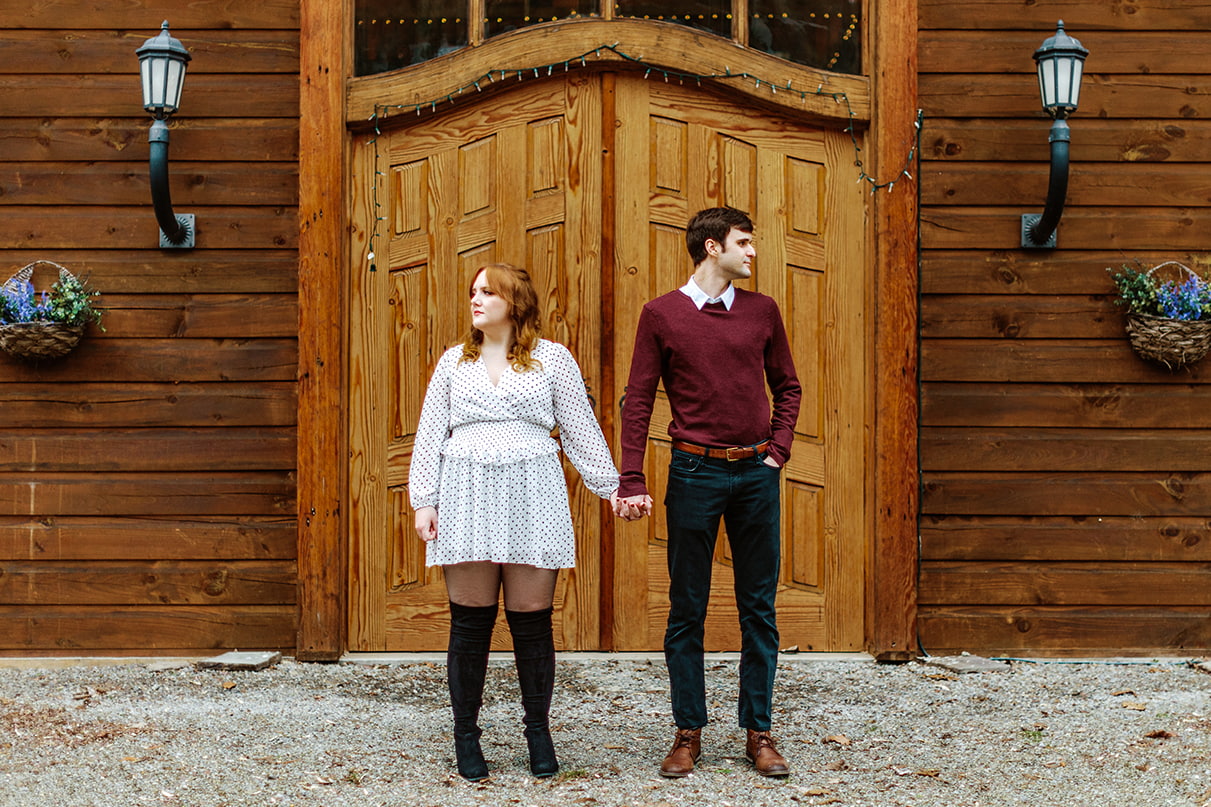 Couple holds hands and looks away from each other while standing in front of wooden doors to a rustic cabin