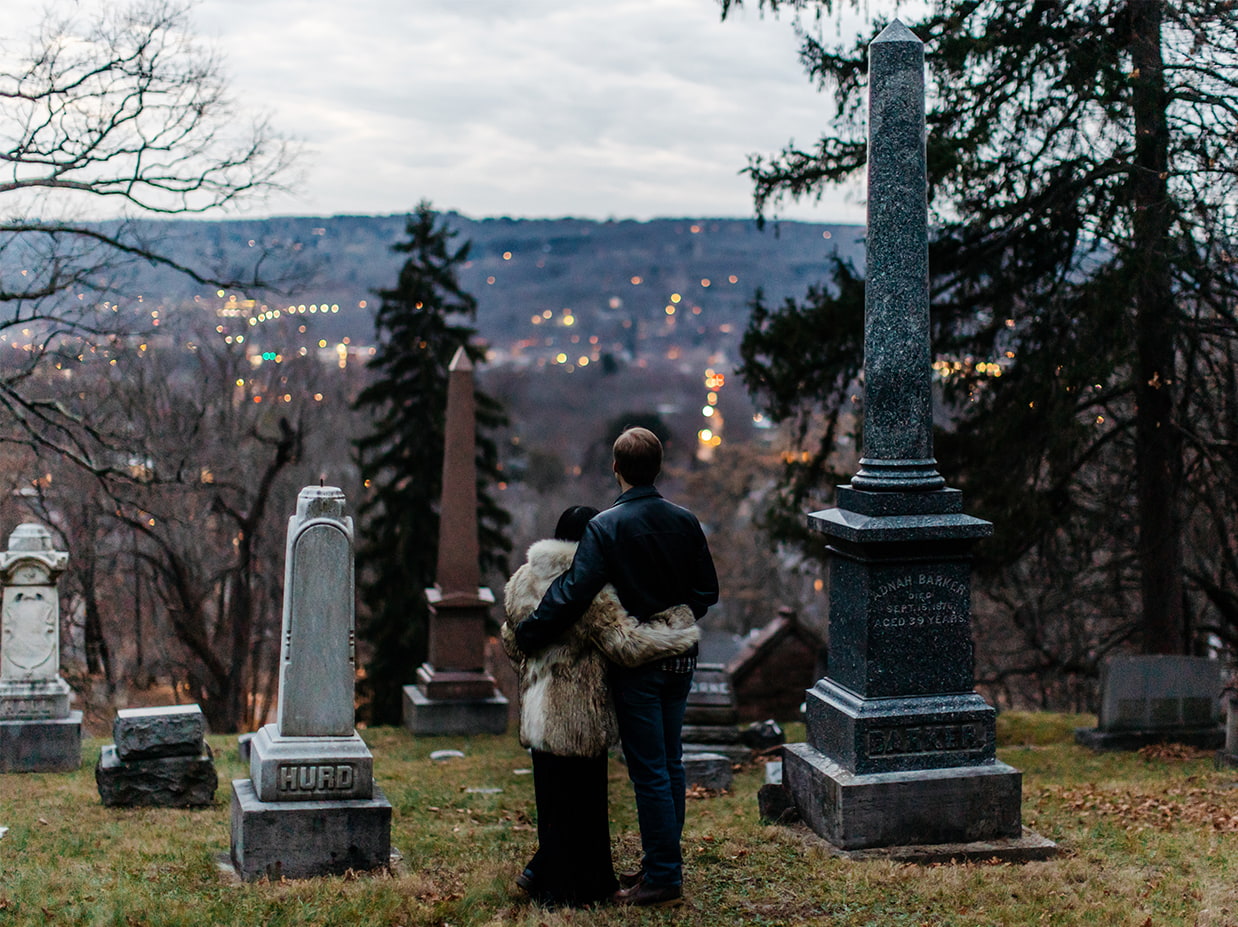 couple looks out into the distance with their arms around each other while standing in a cemetery at night