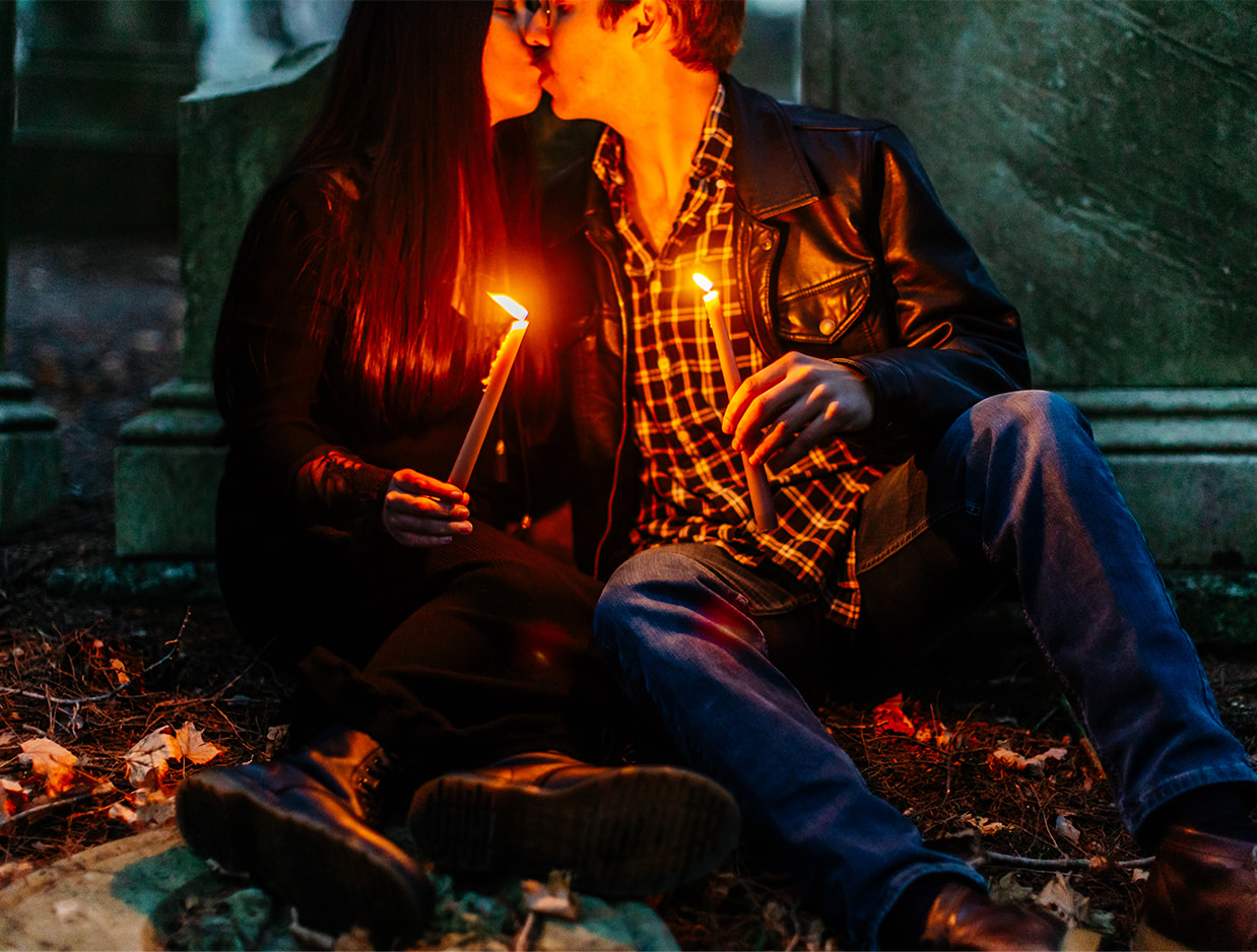 woman in a black dress kisses man in plaid shirt and leather jacket while holding lit candles in a cemetery at night giving a spooky/halloween/stranger things vibe