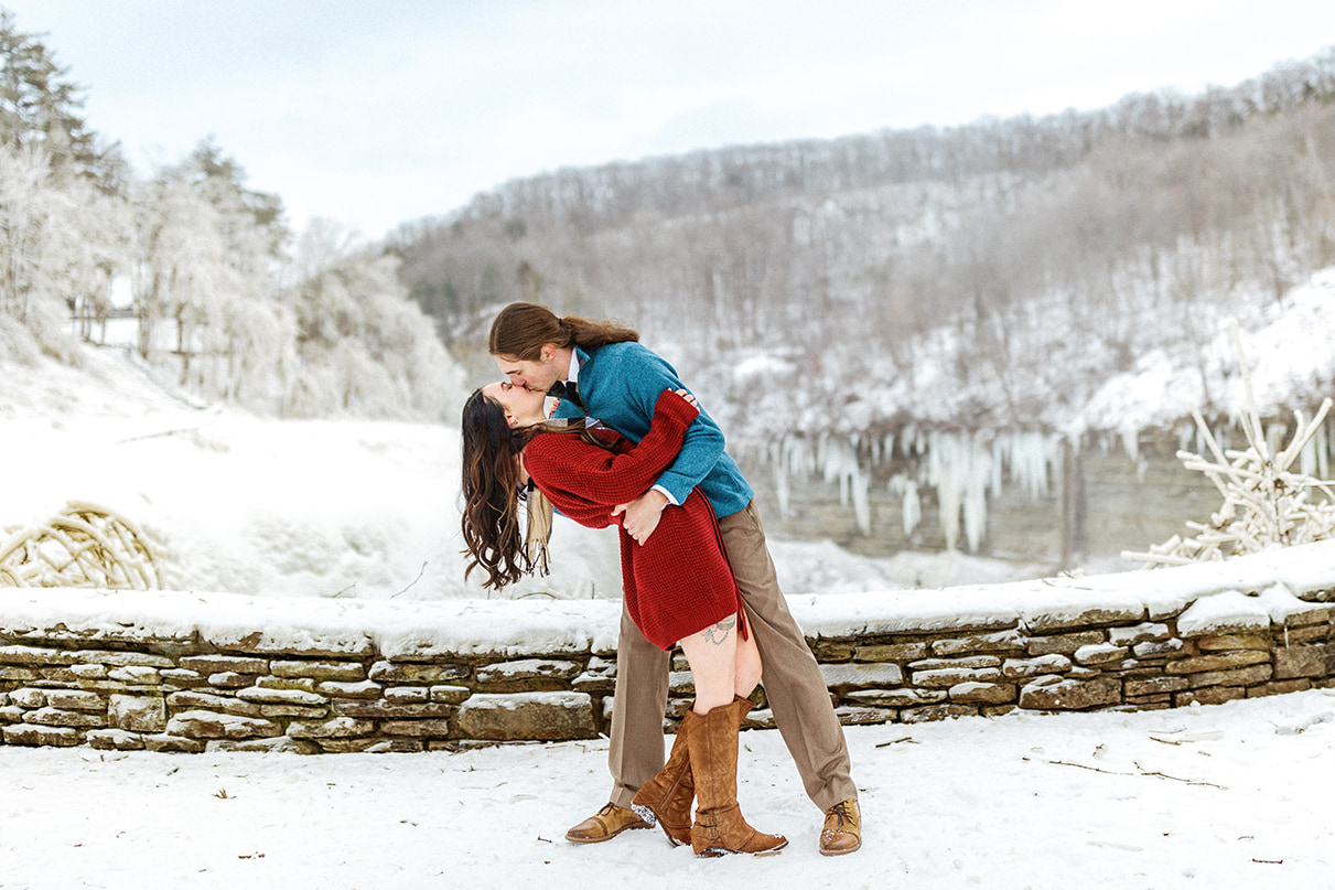 Man in blue sweater dips and kisses fiance during icy outdoor winter engagement photos in Upstate NY