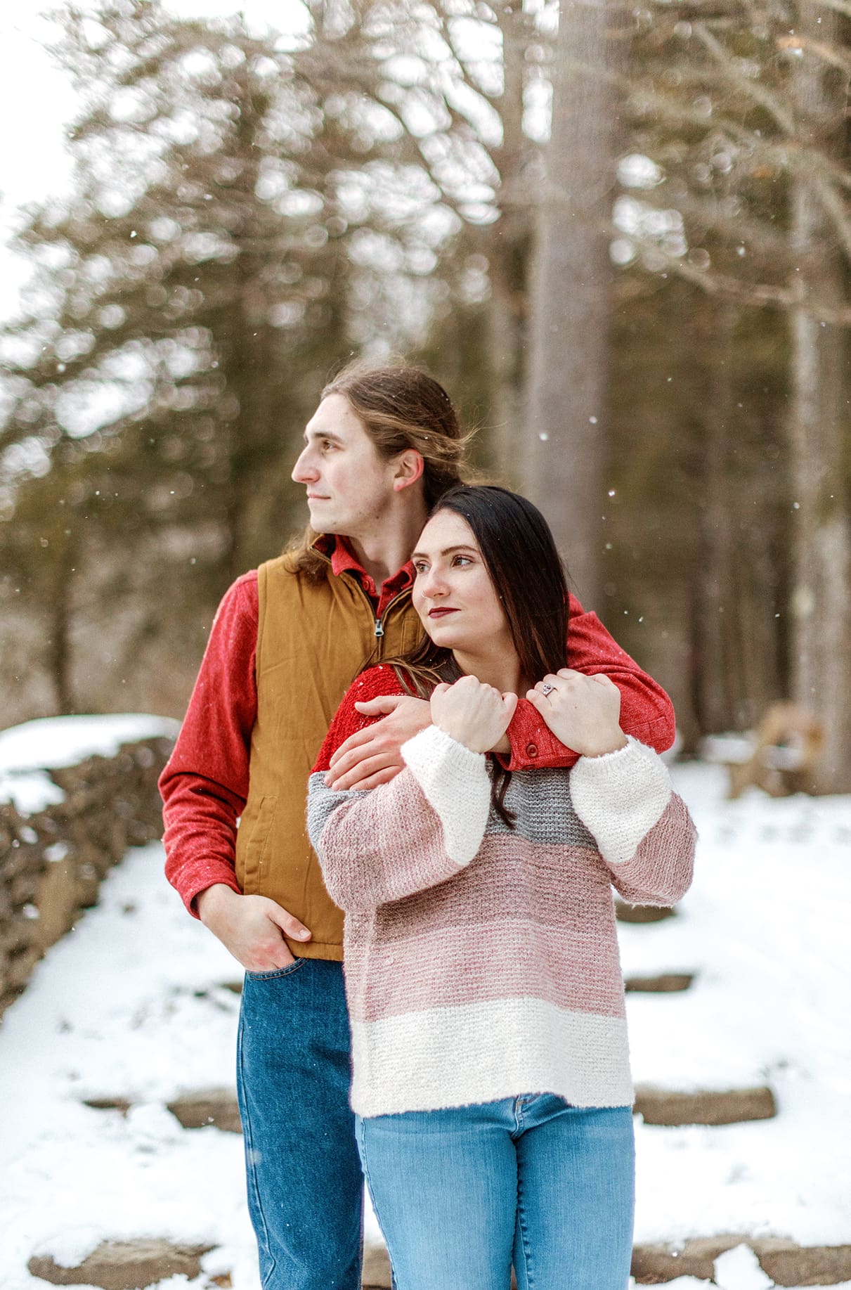 Man hugs woman from behind while they both look into the distance while snow falls during engagement session in Upstate NY