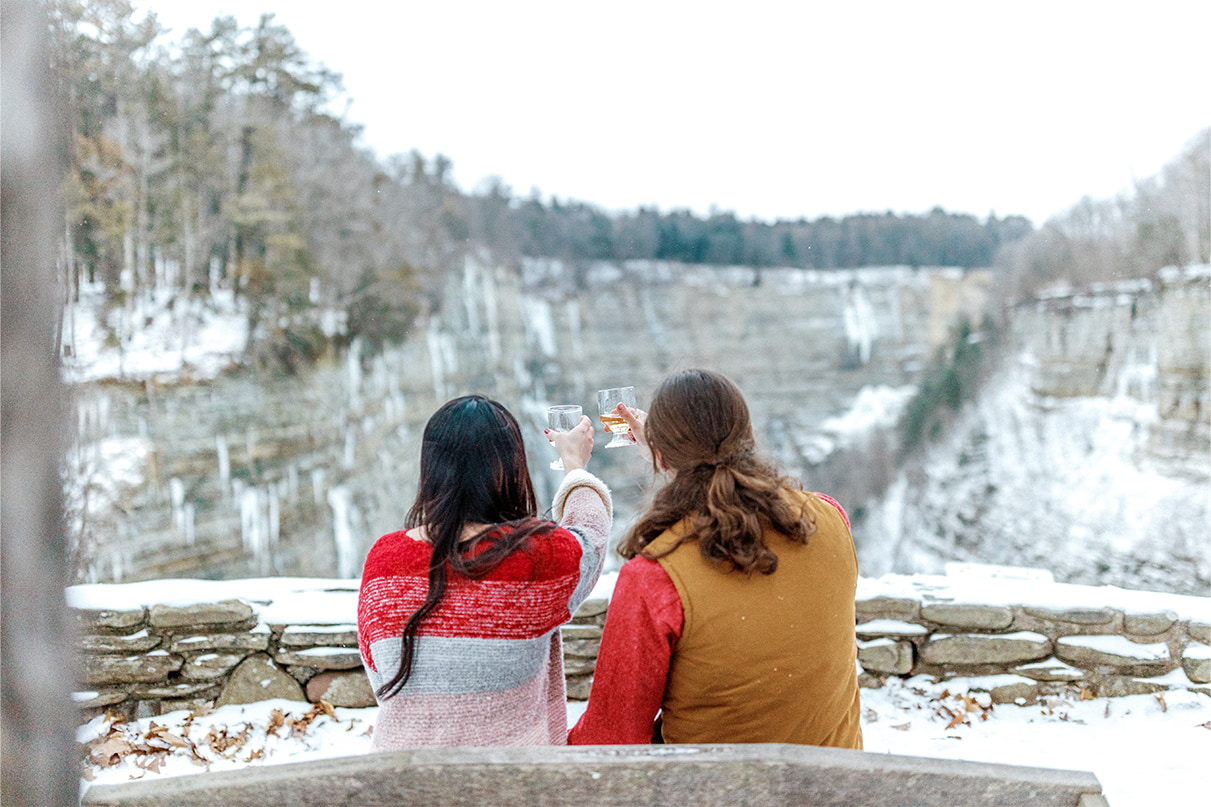 Couple sits on bench and raises two glasses of champagne overlooking snowy gorge in Letchworth State Park