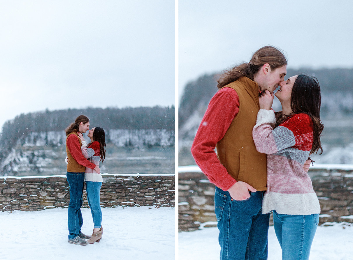 Couple embraces and almost kisses in front of gorge and stone wall while snow falls in Letchworth State Park