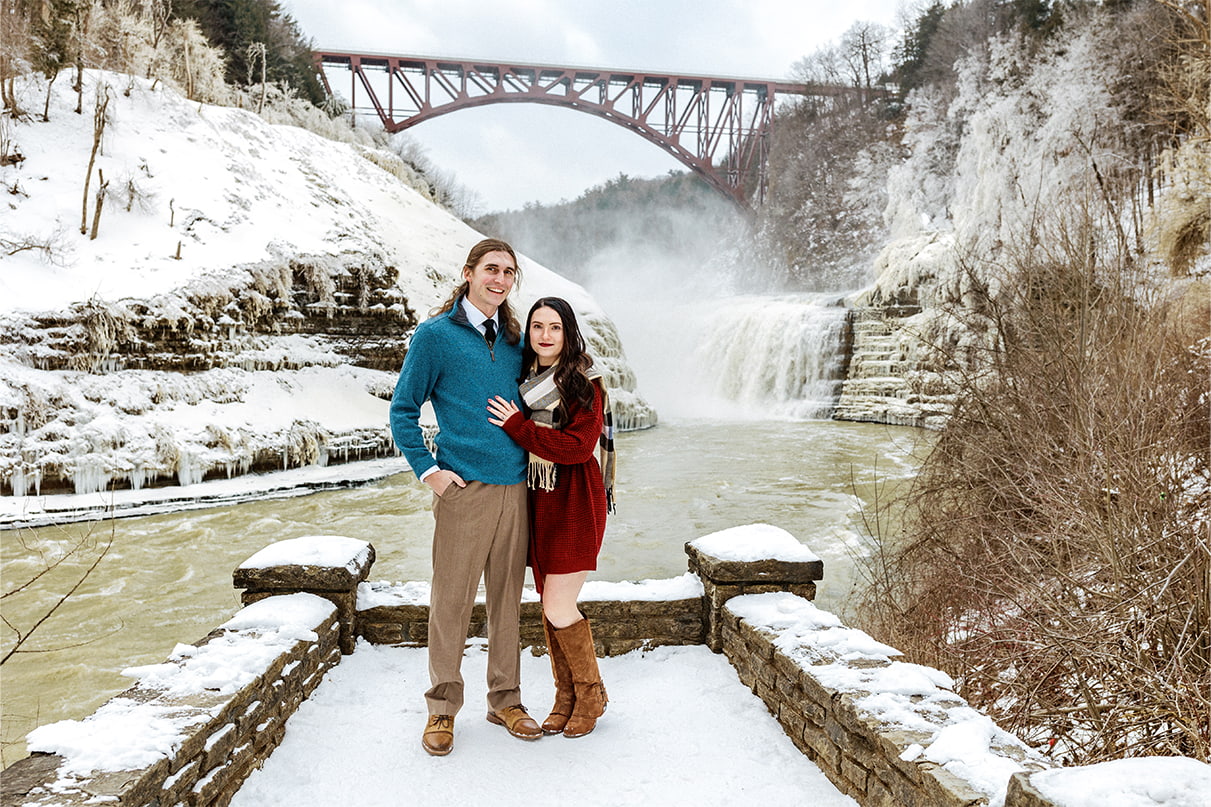 Couple stands and smiles for engagement photos in front of Upper Falls at Letchworth state park
