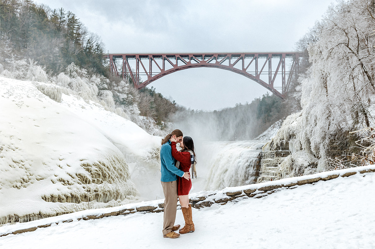 Couple in red dress and blue sweater stands with foreheads together in front of the Upper Falls at Letchworth State Park
