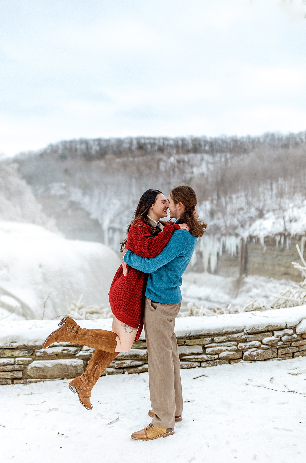 Man in blue sweater lifts woman in red dress in front of icy winter backdrop for upstate ny engagement photos