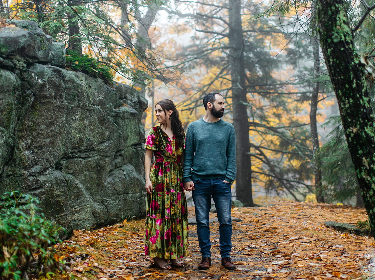 Couple holds hands and looks in opposite directions while standing in front of rocks and fall foliage