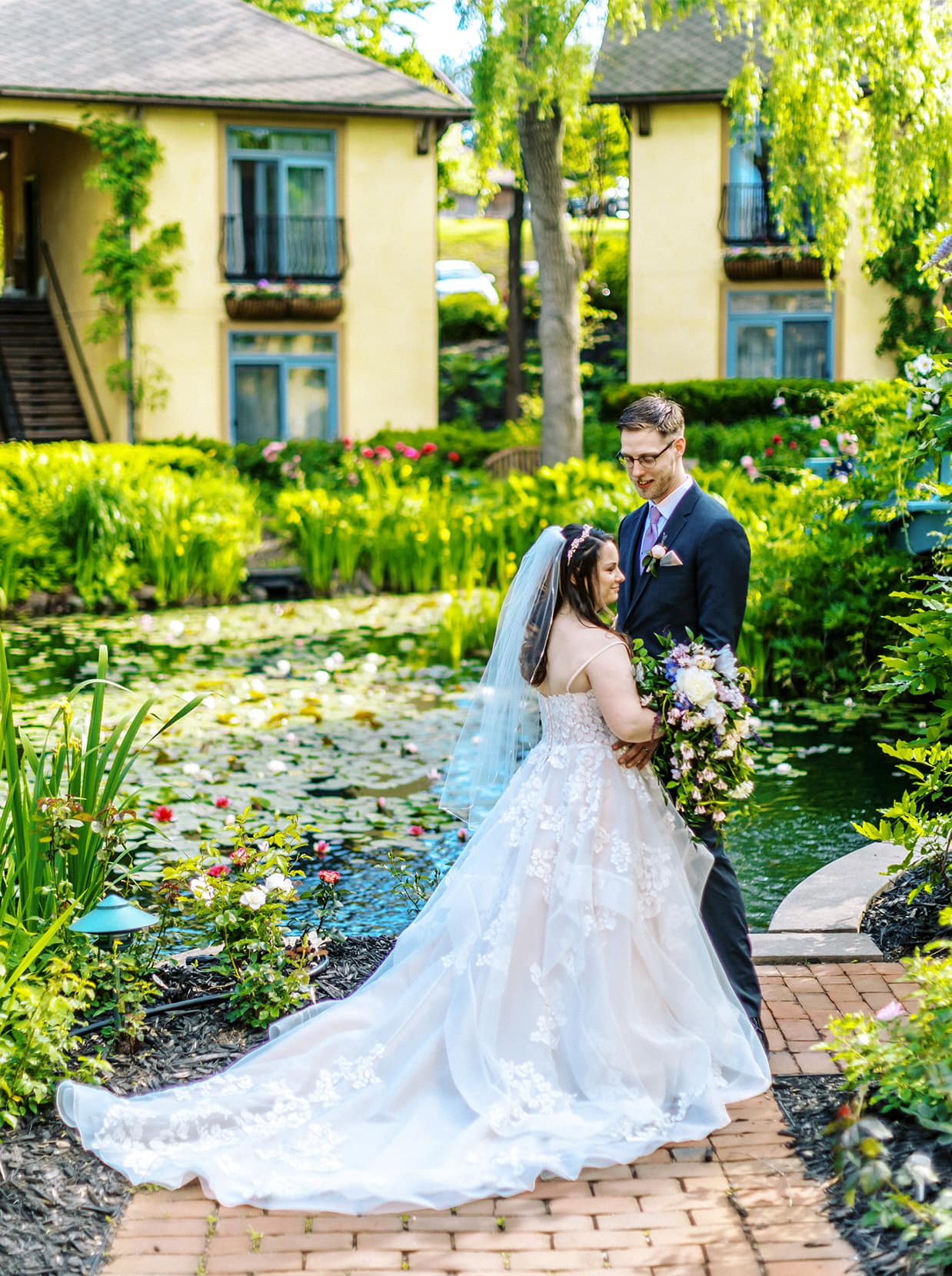 bride and groom stand in lush garden in front of koi pond at mirbeau inn and spa