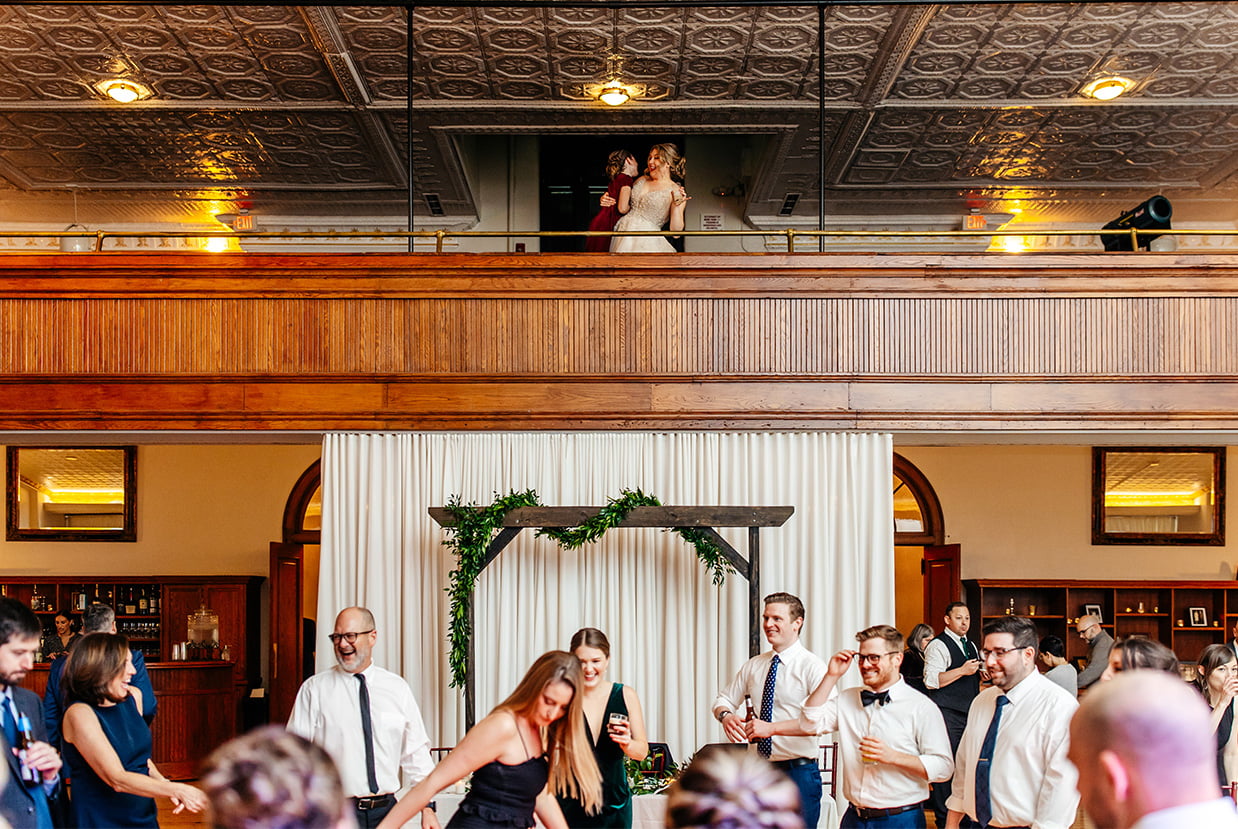 wedding guests dance while bride and flower girl look on from the balcony at The Historic German House in Rochester NY