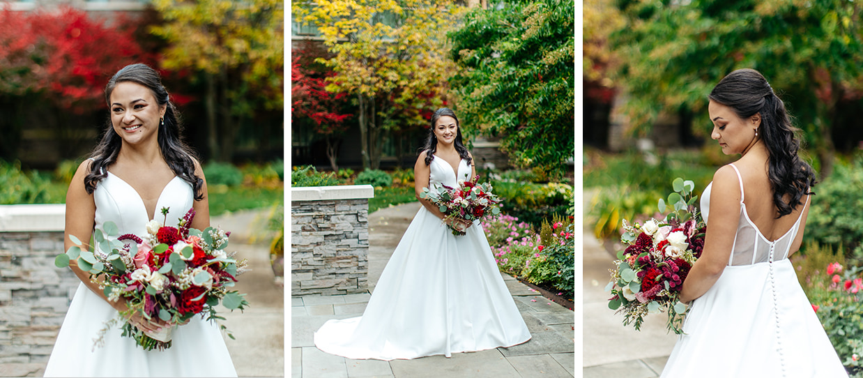 Bride smiles and looks down at her red and pink bouquet in front of colorful trees in a garden at The Lodge at Turning Stone in Verona NY