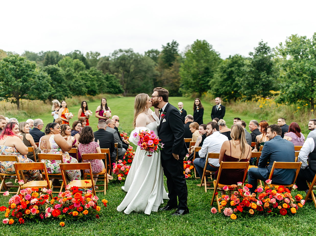 bride and groom kiss at the end of ceremony aisle surrounded by pink, red, and orange flowers at the treman center in newfield NY