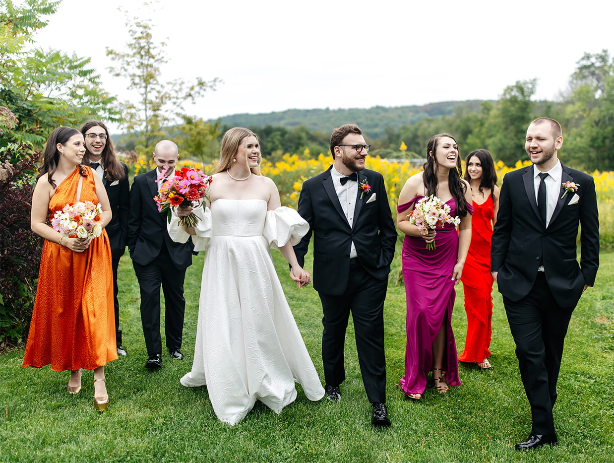 bride, groom, and wedding party wearing colorful attire laugh and walk through a field before their wedding in the finger lakes