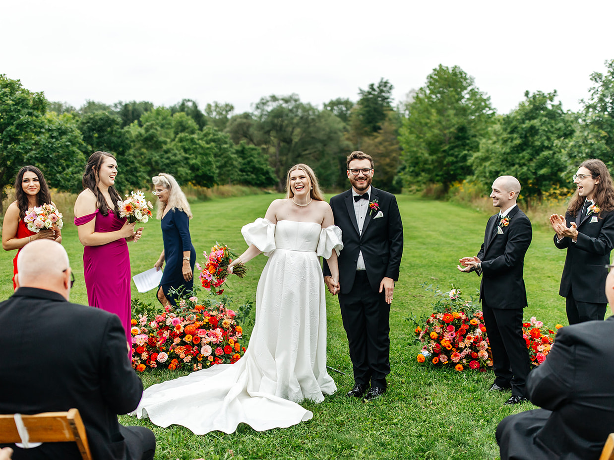 bride and groom stand in front of pink, orange, and red flowers and laugh at the end of their wedding ceremony at the treman center