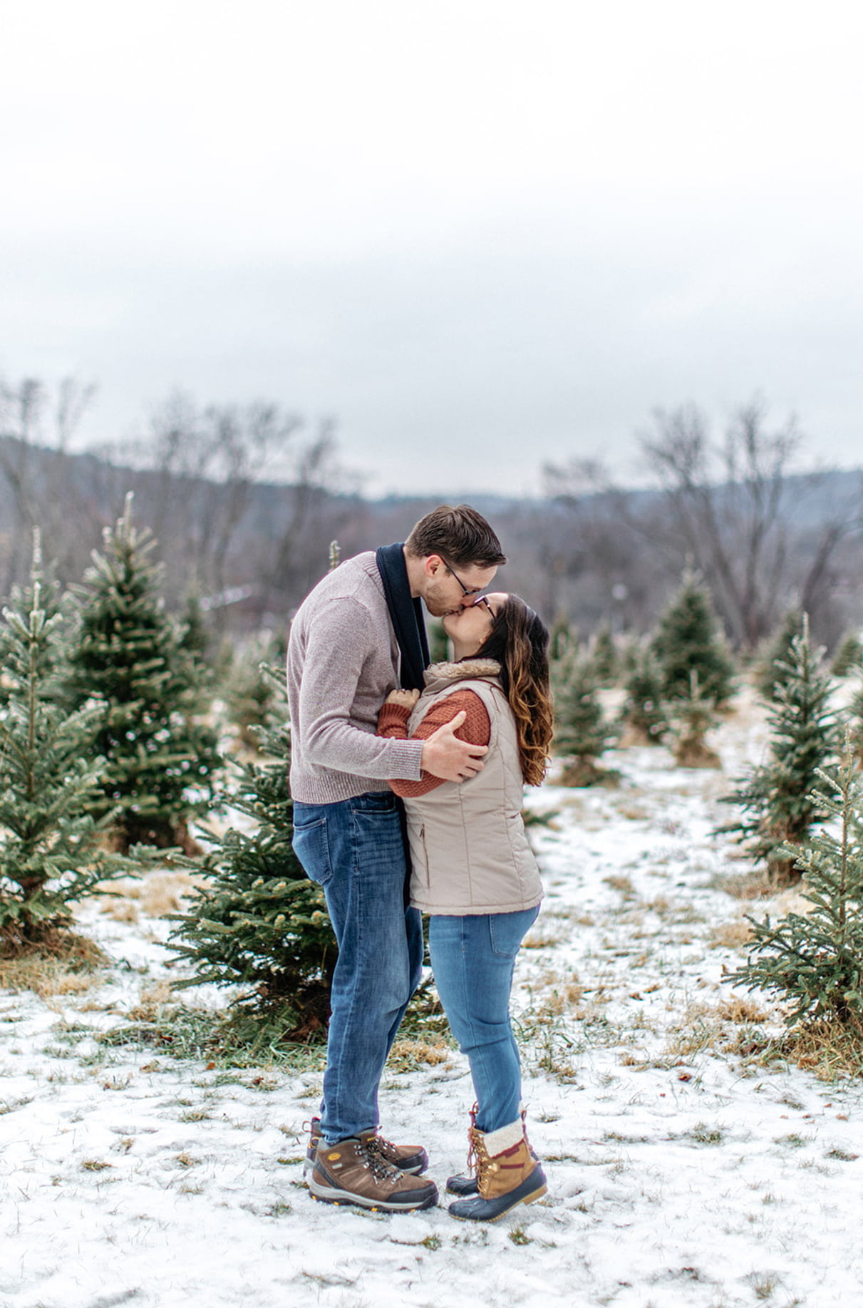 couple stands in snowy field of christmas trees while kissing and woman is holding mans scarf