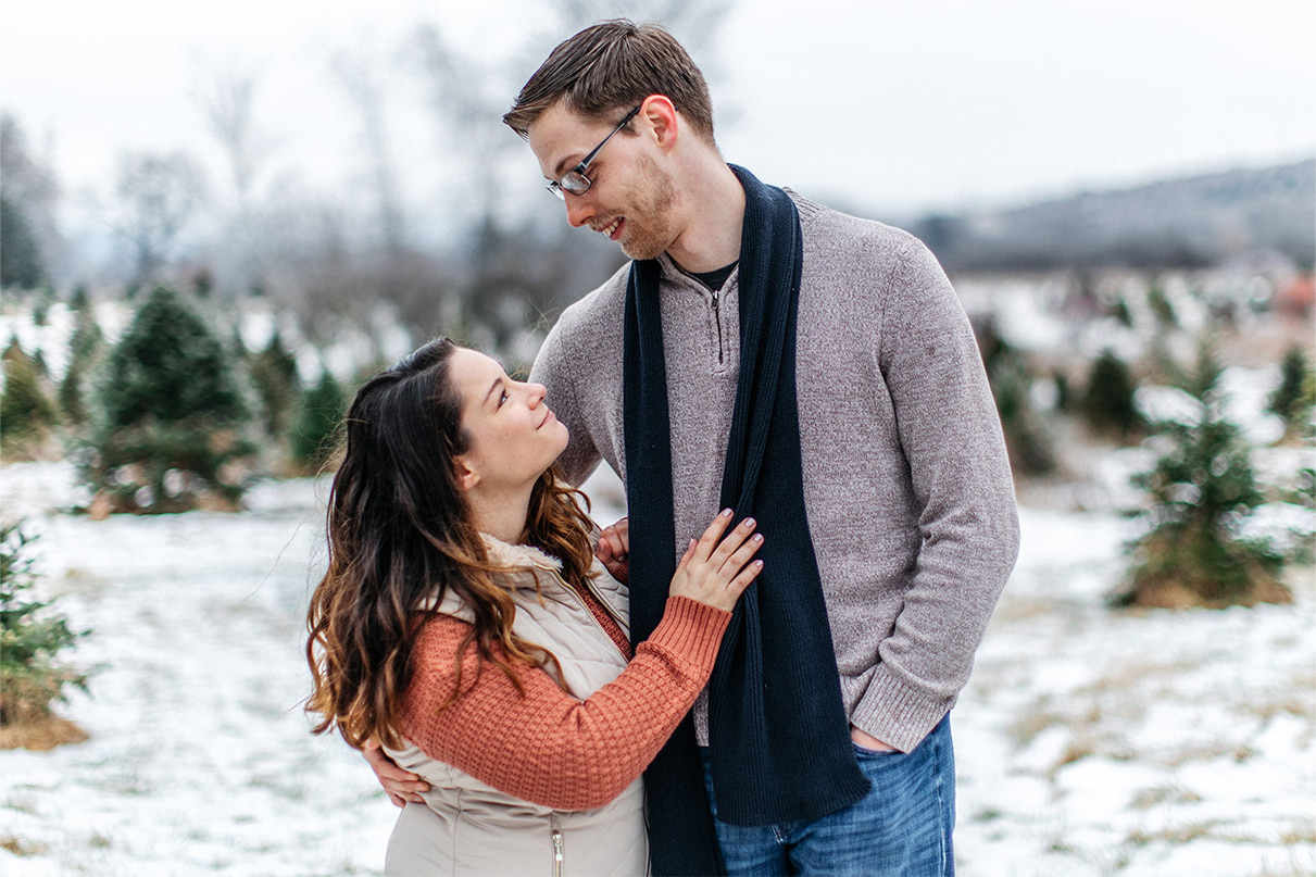 couple smiles at each other while standing in the snowy field of a christmas tree farm in upstate ny