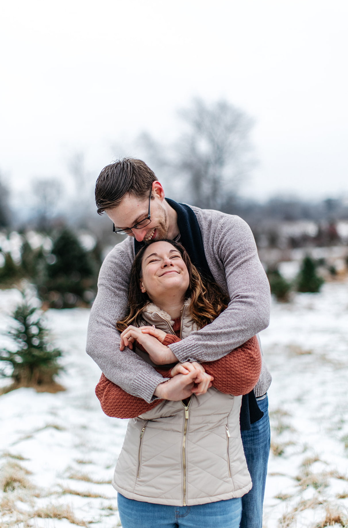 man hugs woman from behind while she smiles and looks up at him on a christmas tree farm in upstate ny