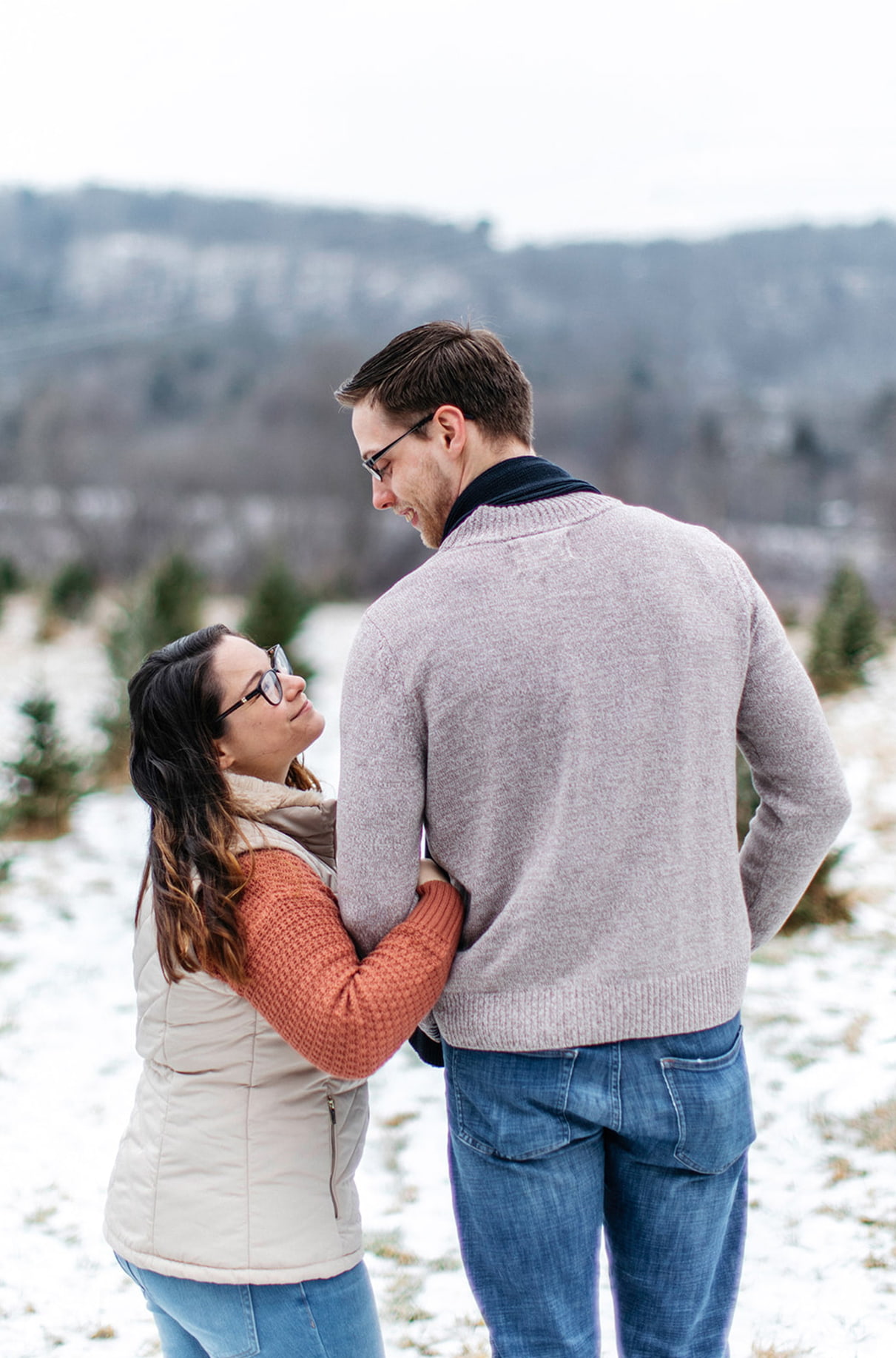couple faces away from camera looking at each other and smiling in a snowy field of a christmas tree farm